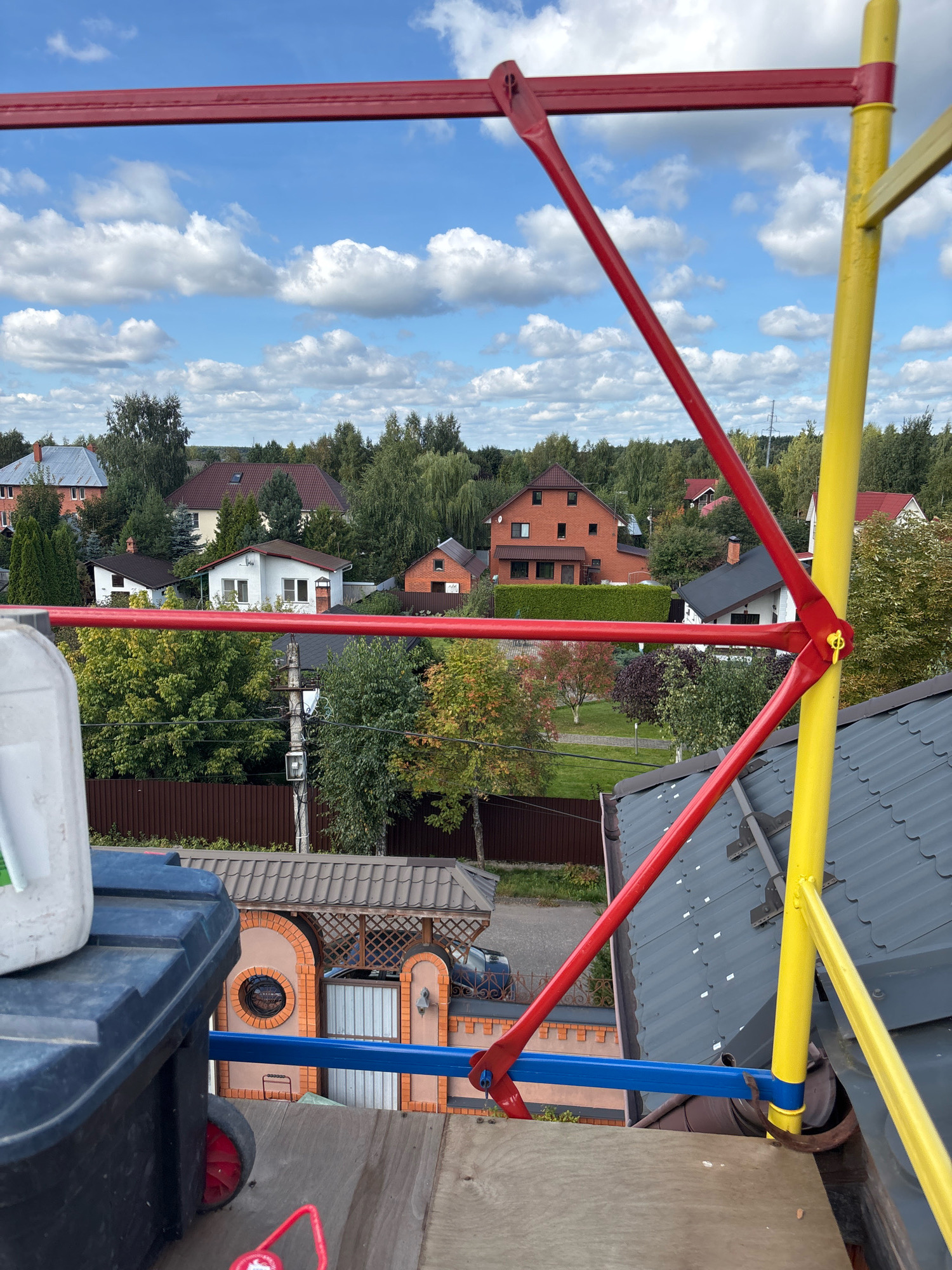 This is a medium-shot, eye-level, outdoor, daytime photograph taken from a high vantage point, likely a rooftop or scaffolding. The foreground is dominated by scaffolding poles, painted red and yellow, with a blue horizontal bar. A dark blue toolbox with red wheels is visible on the left, and a white plastic container sits on top of it. Beyond the scaffolding, a residential neighborhood with houses, trees, and a cloudy blue sky is depicted. The houses have various roof colors, including gray, red, and brown. A brown wooden fence runs through the middle ground. The environment appears to be a suburban or rural area. There are no people visible in the image.