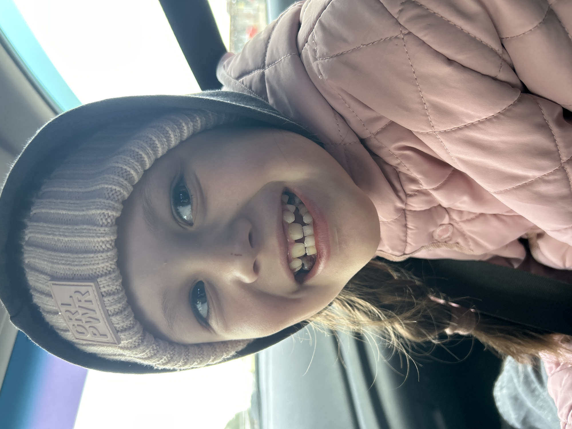 A young girl with fair skin and light brown hair styled in a braid is shown close-up, smiling widely to reveal her teeth. She wears a pink knit hat with a "GRL PWR" patch under a grey hood and a pink quilted jacket. The photograph appears to be taken inside a car, as indicated by the window and dark interior elements in the background. The exact location within the city of Chaikovsky, Russia cannot be determined from the image alone. The subject's action is smiling.