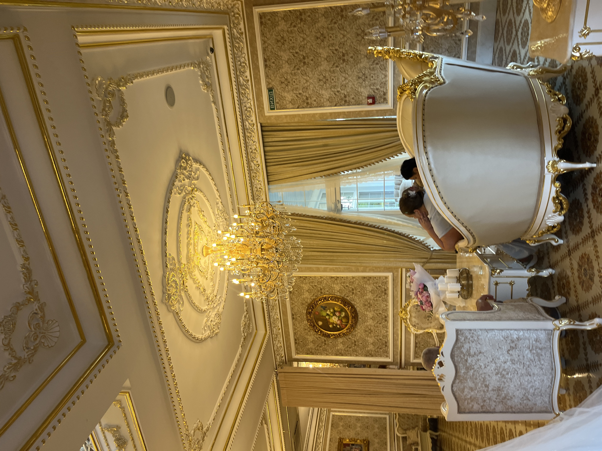 The image depicts an opulent interior scene, likely a dining room or lounge, in Dubivtsi, Ukraine. The room is adorned with intricate gold detailing on the ceiling, walls, and furniture, suggesting a lavish and perhaps historical setting. A large, multi-tiered crystal chandelier hangs from the ornate ceiling, illuminating the room.

In the foreground, two individuals are seated at a polished table. The person on the left is facing away from the camera, with their head slightly turned. The person on the right is partially visible, with their arm resting on the back of a plush, cream-colored armchair. A bouquet of pink flowers and several white candles are arranged on the table between them.

The furniture is elaborate, with carved gold accents and upholstered in light-colored fabrics. The walls are covered in a textured wallpaper with a gold damask pattern, bordered by gold trim. Heavy, gold-colored curtains frame a large window to the left of the scene, which appears to look out onto a garden or outdoor area. A framed oval painting of a floral arrangement hangs on the wall between the window and the table.

To the right of the main seating area, a sign with the word "ВИХІД" (meaning "EXIT" in Ukrainian) is visible on the wall, indicating an emergency exit. The floor is covered in a patterned carpet with intricate designs in shades of brown and gold. The overall impression is one of luxury and grandeur, with meticulous attention to decorative detail.