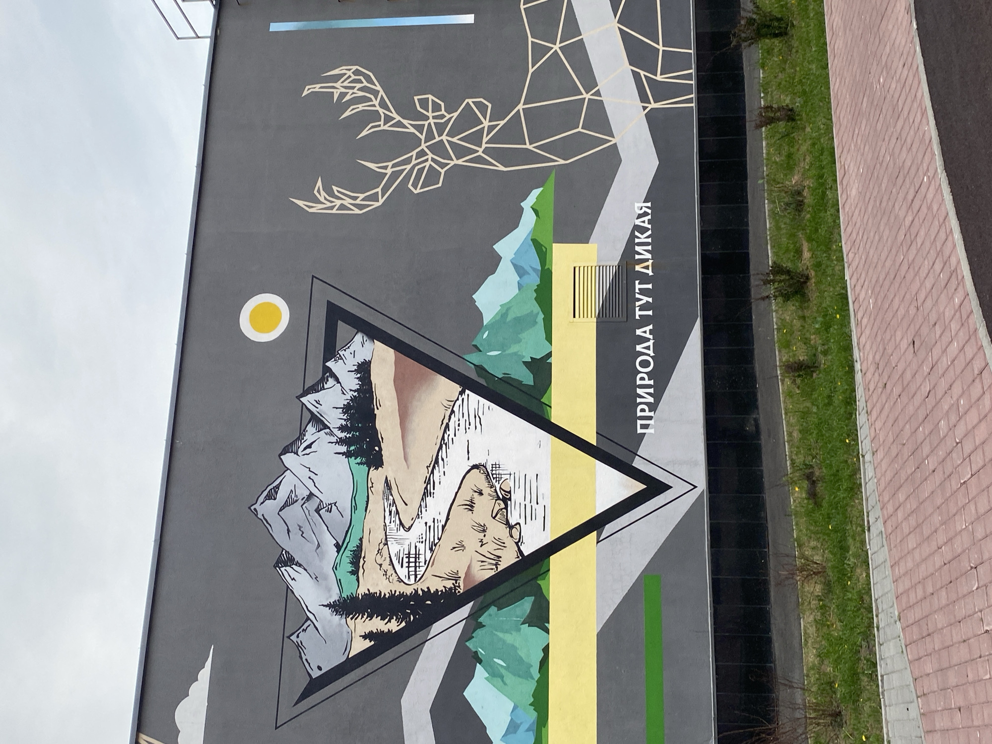 Here is a description of the image:
This is a low-angle, color photograph of a mural on the side of a building. The mural features a geometric design, including a stylized set of antlers at the top, and a triangular design containing a landscape with mountains, trees, and a river. A yellow circle represents the sun. To the right of the triangular design, Cyrillic text reads "ПРИРОДА ТУТ ДИКАЯ". The mural is painted on a gray wall, with additional geometric shapes and colored stripes extending around the central landscape.
The building is situated next to a brick-paved area and a narrow strip of green grass and small shrubs. The sky is overcast. The photograph suggests the location may be the city of Yekaterinburg, Russia, based on the Cyrillic text.