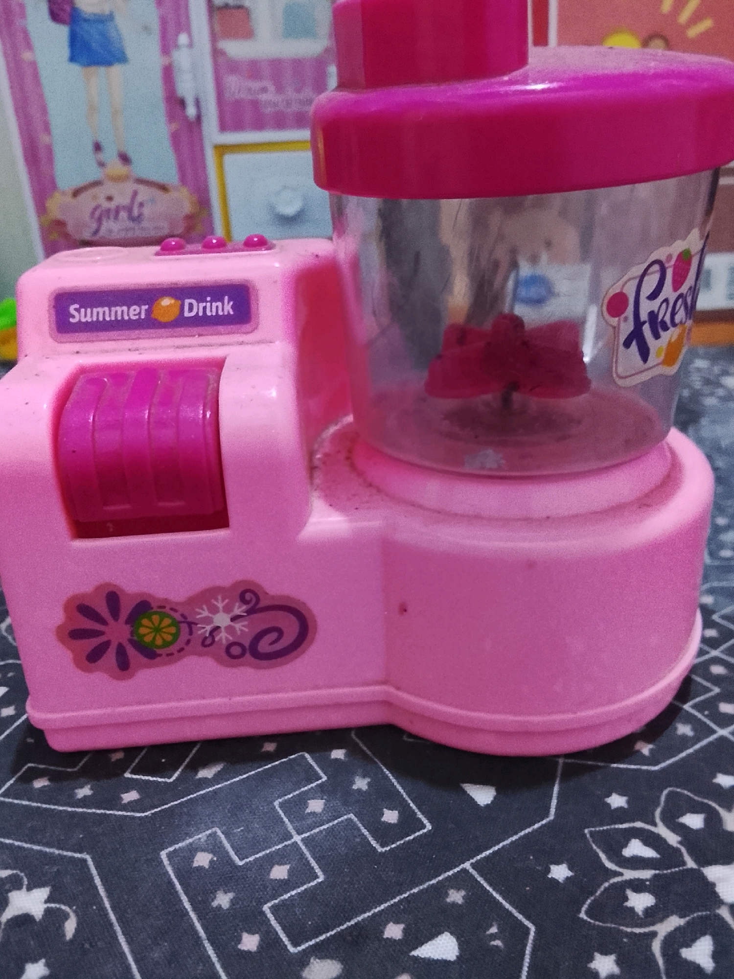 The image shows a pink toy blender. The blender has a transparent cup with a pink lid and a pink base. On the side of the blender, there is a purple label that says "Summer Drink" with an orange circle in between. There are three pink buttons on top of the blender. The blender is decorated with stickers of flowers and snowflakes. The background of the image is a patterned fabric. The image does not provide any information about the city of Ujungrusi, Indonesia.