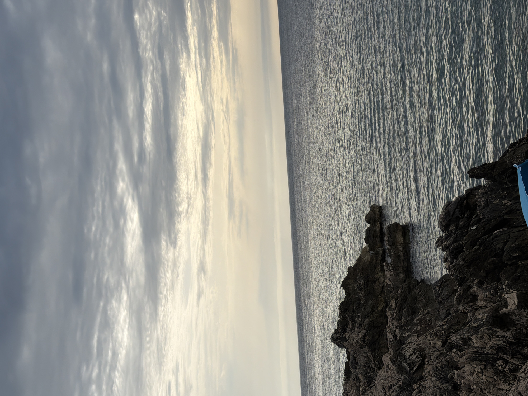 The image shows a rocky shoreline with the Adriatic Sea stretching out to the horizon under a cloudy sky. The foreground is dominated by dark, jagged rocks, with the water lapping at their base. A fishing rod is visible, propped against the rocks. In the background, the sea meets the sky in a soft gradient of muted colors. The scene suggests a tranquil, perhaps overcast, day at the coast. The location is identified as Kunje, Montenegro.