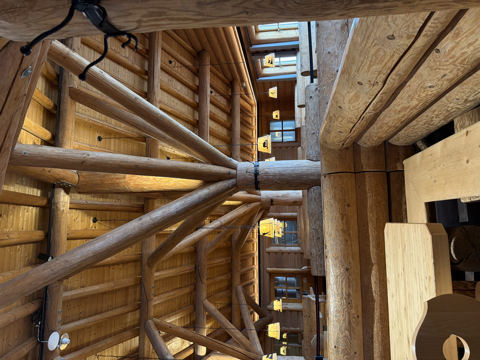 This is an interior shot of a large, wooden building, possibly a lodge or resort, in an unknown location. The architecture features extensive use of logs and timber in the construction. The image is taken from a low angle, looking up at the complex network of wooden beams and structural elements. The ceiling is visible through gaps in the construction, with skylights and hanging lamps. There is no human presence or activity explicitly visible.