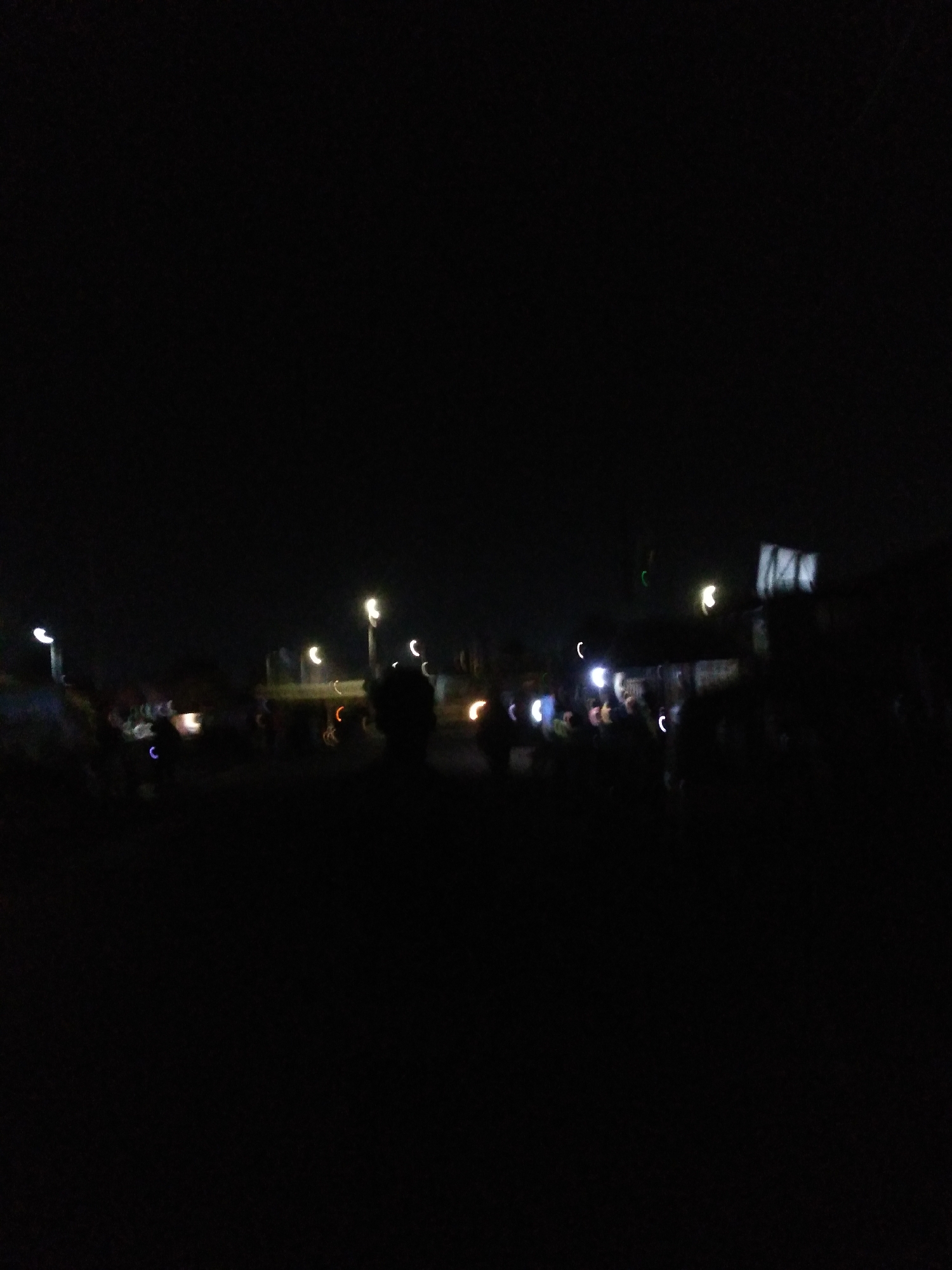 The image depicts a nighttime scene in what appears to be an outdoor market or gathering area in Jos, Nigeria. The overall lighting is dim, with artificial lights creating blurry streaks and circles of illumination against the dark background. Several sources of light are visible, including streetlights and possibly lights from stalls or vehicles. There are indistinct human figures scattered throughout the scene, suggesting the presence of people engaged in various activities, though their specific actions are not discernible due to the low light and motion blur. Some of the light sources appear to be associated with vehicles or handheld lights. The environment suggests an urban setting, with the lights hinting at infrastructure like roads or buildings, consistent with a location in Jos.
