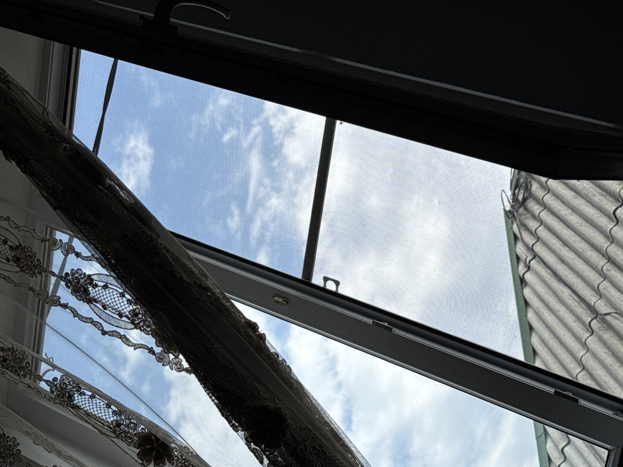 The image shows a view looking up and out of a window with a screen and curtains. The sky is visible with some clouds. A portion of a tiled roof is visible below the window. The scene is not in a city; it is a view from what appears to be a building with a roof. There are no people or discernible actions taking place. The location of Tsiyab-Tsoloda, Russia is irrelevant to the visual content of the image.