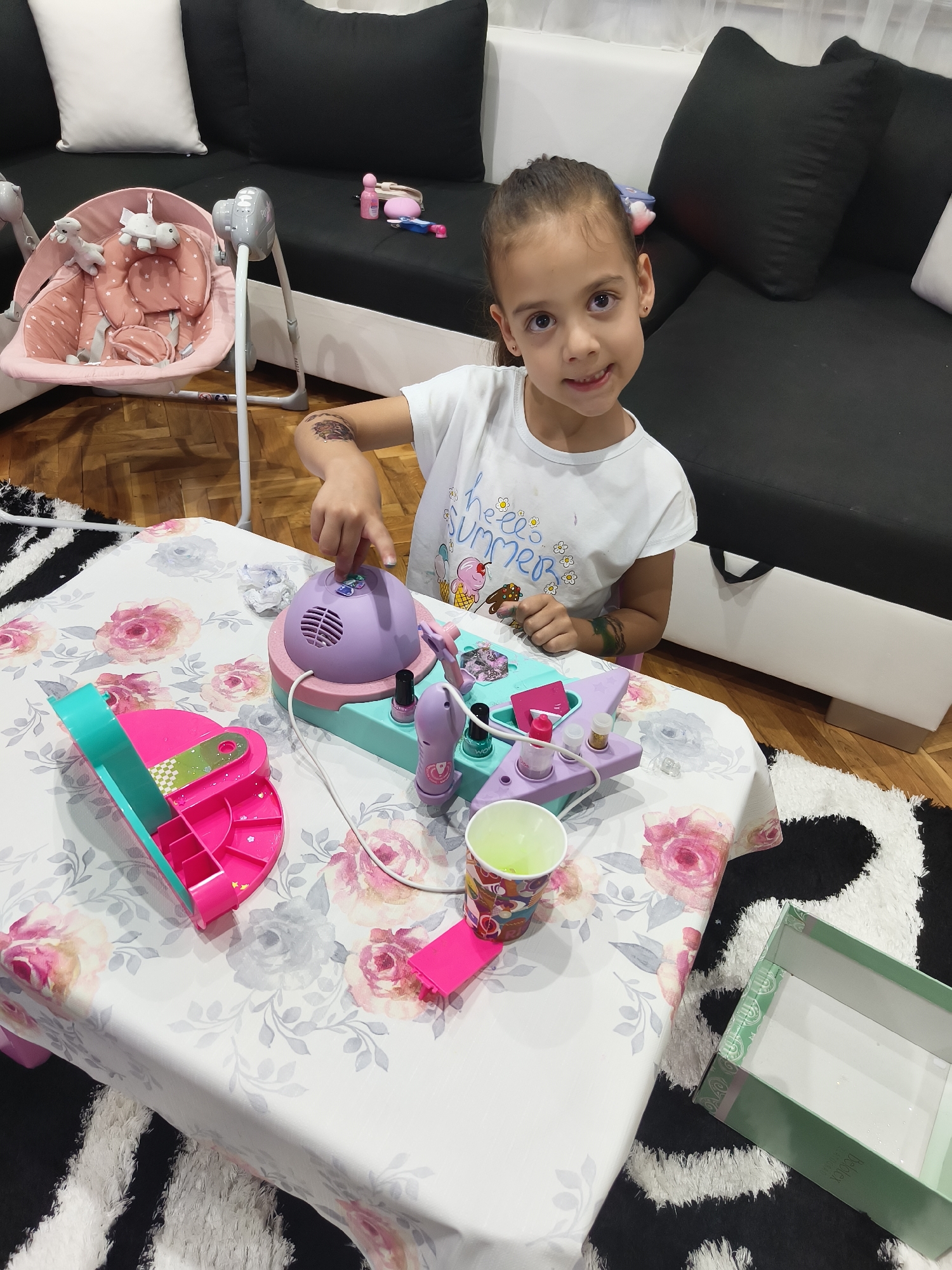 A young girl is captured indoors, likely in a living room or play area, engrossed in playtime with a toy nail salon set. She is seated at a white table adorned with a floral tablecloth featuring pink and grey roses. The girl, appearing to be of primary school age, has dark hair tied back and wears a white t-shirt with the visible text "hello summer" accompanied by ice cream cones and flowers. A temporary tattoo is noticeable on her right upper arm. She is looking directly at the camera with a gentle smile, suggesting a cheerful mood.
On the table in front of her rests a pink and purple toy nail salon set, which includes a small "nail dryer" unit, toy nail polish bottles (one with "WOW" text visible), and other accessories. Her right hand is positioned near the dryer, indicating active engagement with the toy. To her left, an open pink and teal container, possibly part of the toy set, is on the table. A colorful paper cup holding a light green liquid and a crumpled piece of paper are also present on the table surface.
In the background, a pink baby bouncer or swing, decorated with star patterns and holding plush unicorn toys, is visible. Behind the bouncer, a black sofa with black and white cushions is arranged, with small pink items placed on it. The flooring beneath the rug is wooden, and a black and white patterned rug partially covers it. A green box, partially showing the brand name "Bolden," is on the floor to the right of the table. The lighting suggests it is daytime. This scene depicts a domestic setting of childhood play in Svilengrad, Bulgaria.