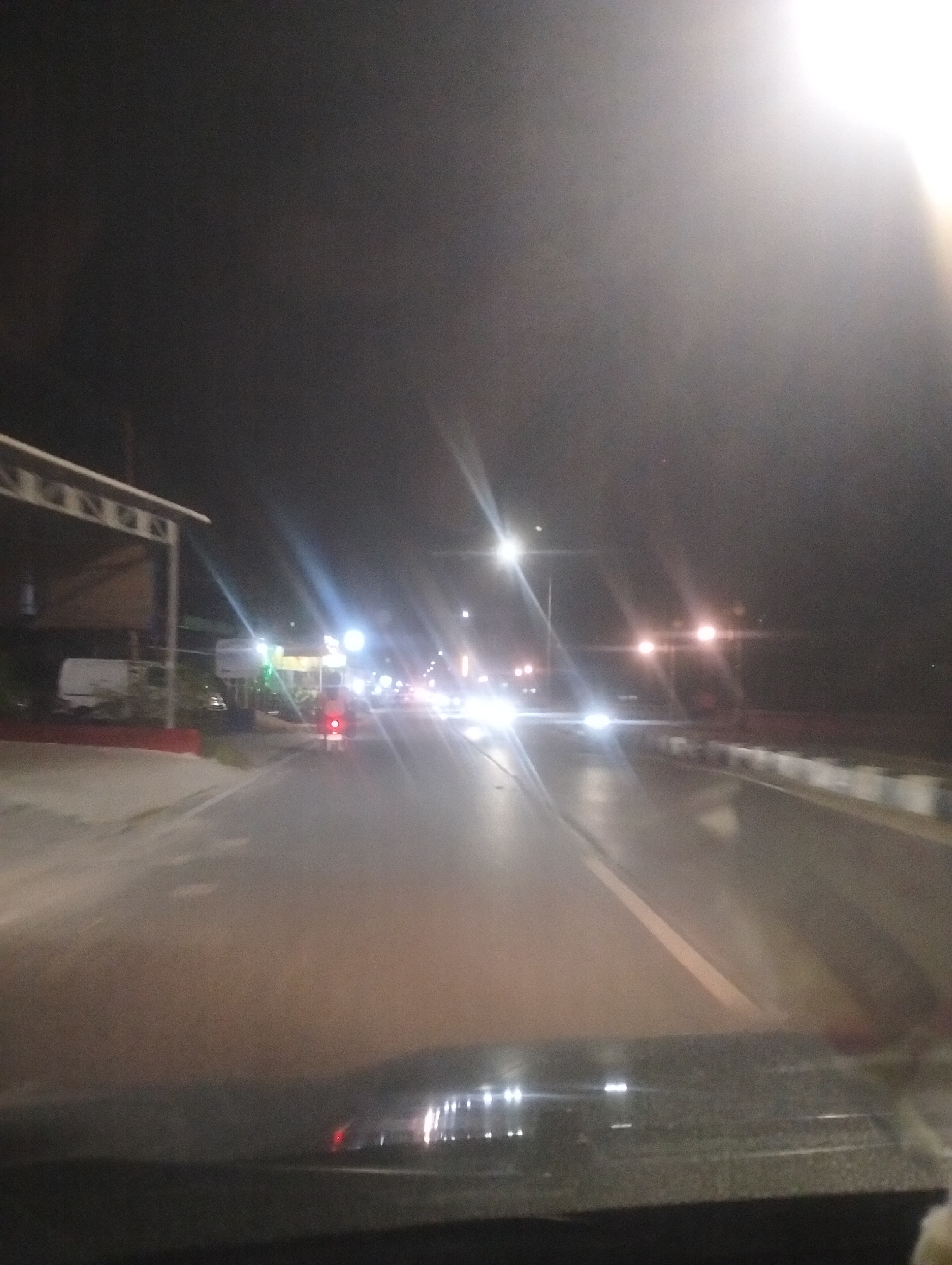 The image depicts a night scene on a road in Dumai City, Indonesia. The perspective is from inside a vehicle, looking forward. Streetlights illuminate the road, creating streaks of light due to the low light conditions and possibly motion. Various vehicles, including a motorcycle with a red taillight and other cars, are visible on the road. On the left side of the road, there appears to be a structure with a canopy and some parked vehicles, possibly a roadside establishment or parking area. The road itself has white lane markings. The overall impression is of a typical night drive through an urban or semi-urban area in Dumai City.