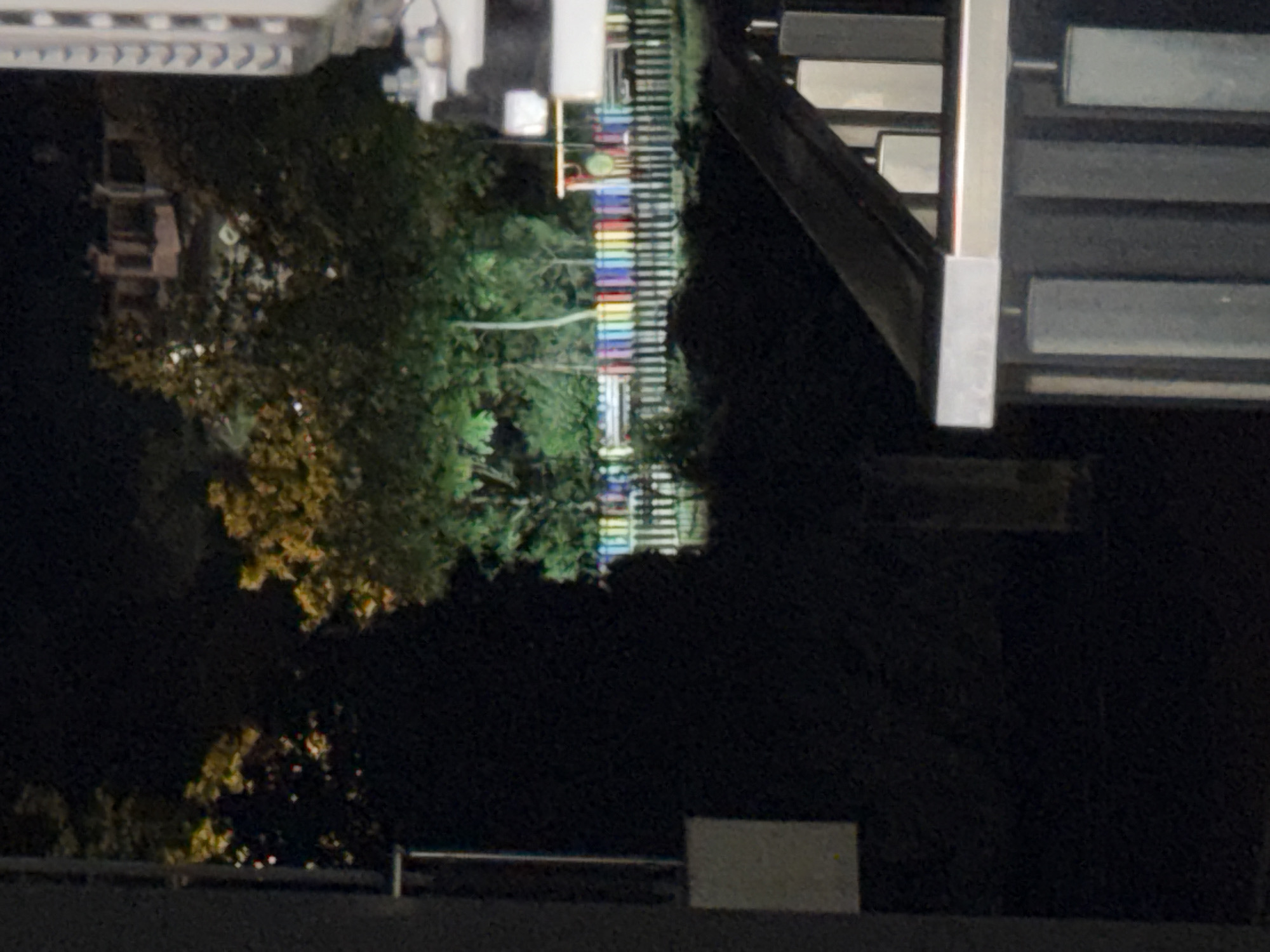 An outdoor nighttime shot captures a scene that appears to be a park or recreational area. In the foreground, a dark, diagonal railing or barrier suggests a viewing platform or walkway. Behind this, a wooden fence with a colorful top railing, painted in alternating sections of red, yellow, blue, and green, stretches across the middle ground. Beyond the fence, trees are illuminated by a bright light source, casting green hues on the foliage. A building with multiple windows is visible in the background, and a tall, multi-story structure with a textured facade stands on the far right. The overall ambiance is one of a dimly lit urban environment at night.