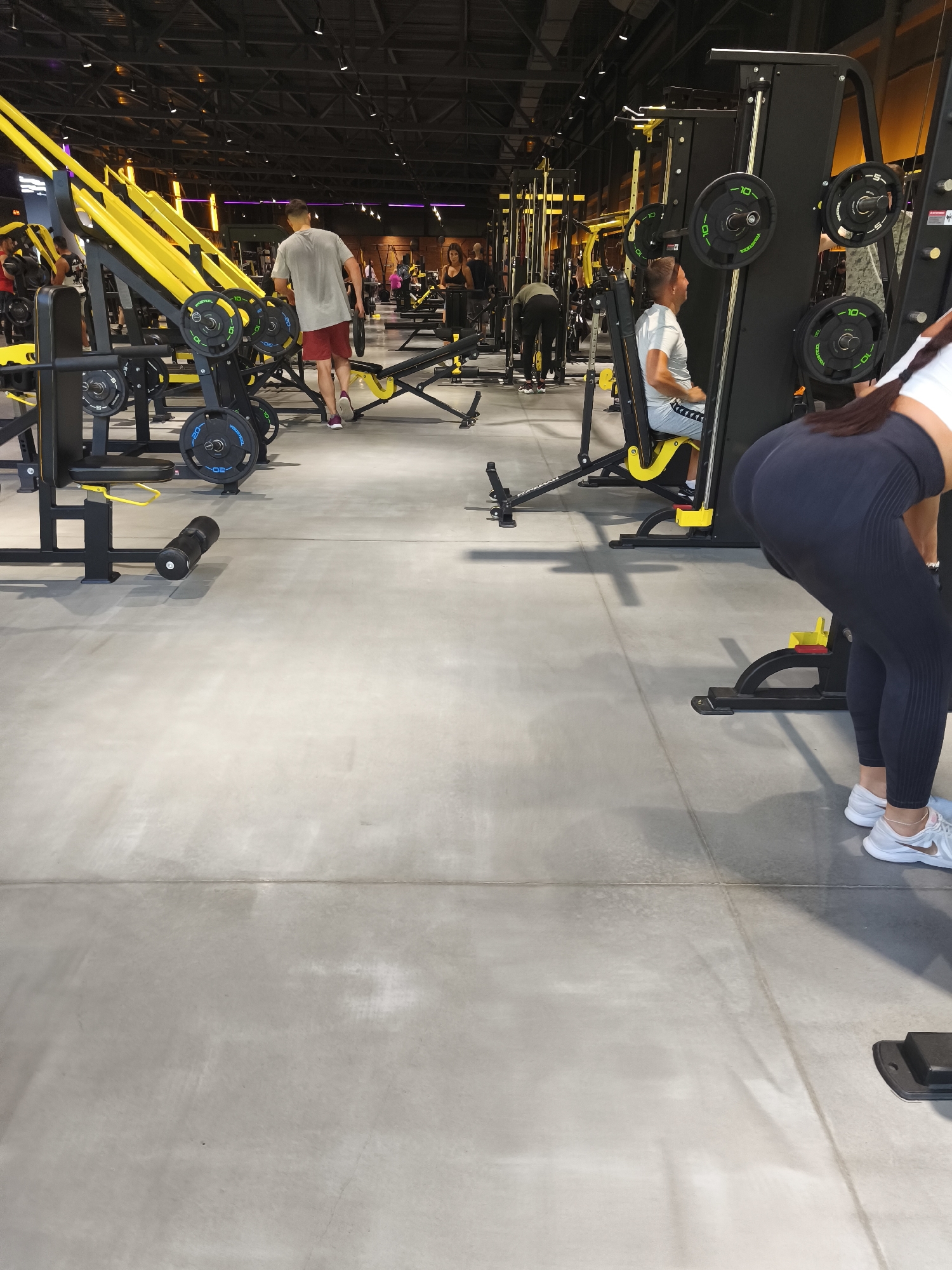 The image depicts the interior of a modern gym, likely located in Komsomolsk-on-Amur, Russia, given the context. The gym features a variety of weight training equipment, primarily in yellow and black, with weights stacked on some machines. The floor is a smooth, light gray concrete.
In the foreground, a person wearing dark leggings and white athletic shoes is bent over, with their back to the camera. Their hair is dark and long, with a braid visible. They appear to be engaged in some form of exercise.
Further back, a man in a light gray t-shirt and red shorts is walking away from the camera, holding a weight plate in his right hand. He is near a set of yellow exercise machines. To his left, another man in a white t-shirt and gray shorts is seated on a weight machine, facing away from the camera.
Other individuals are visible in the background, engaged in various activities within the gym environment. The lighting suggests an indoor setting with artificial illumination. The overall impression is one of a functional and well-equipped fitness facility.