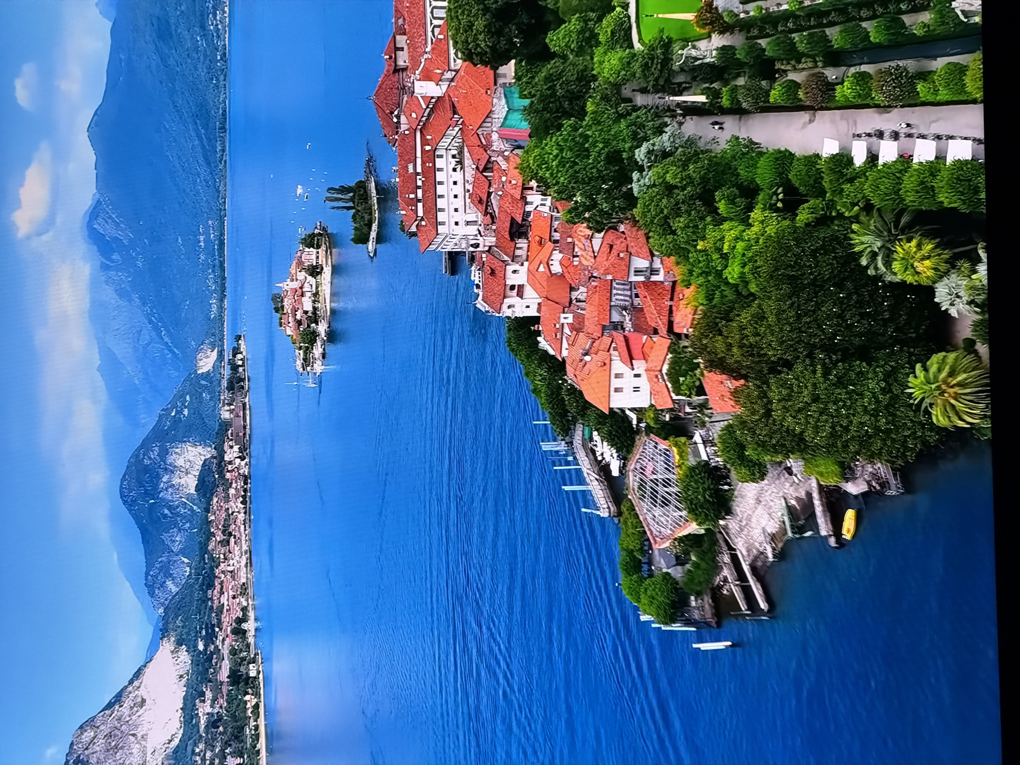 The image depicts a scenic aerial view of a coastal town nestled beside a large, vibrant blue body of water. In the distance, majestic mountains with rugged, rocky peaks rise against a clear sky with scattered clouds. A small island with a cluster of buildings and greenery sits in the water, further out from the mainland.
The foreground showcases a town with numerous buildings featuring red tile roofs, characteristic of Mediterranean architecture. These buildings are densely packed along the coastline, interspersed with lush green trees and gardens. A pathway lined with manicured hedges and trees winds through the town, with a few figures visible walking along it, suggesting a public space or park.
The water is calm with gentle ripples, indicating a serene environment. A small yellow boat is moored near the shore, and several piers extend into the water. The overall impression is one of a tranquil and picturesque location, possibly a tourist destination given the well-maintained gardens and pathways.
It is important to note that this scene is not located in Chirchik, Uzbekistan. The architecture, landscape, and geographical features are inconsistent with the known characteristics of Chirchik.