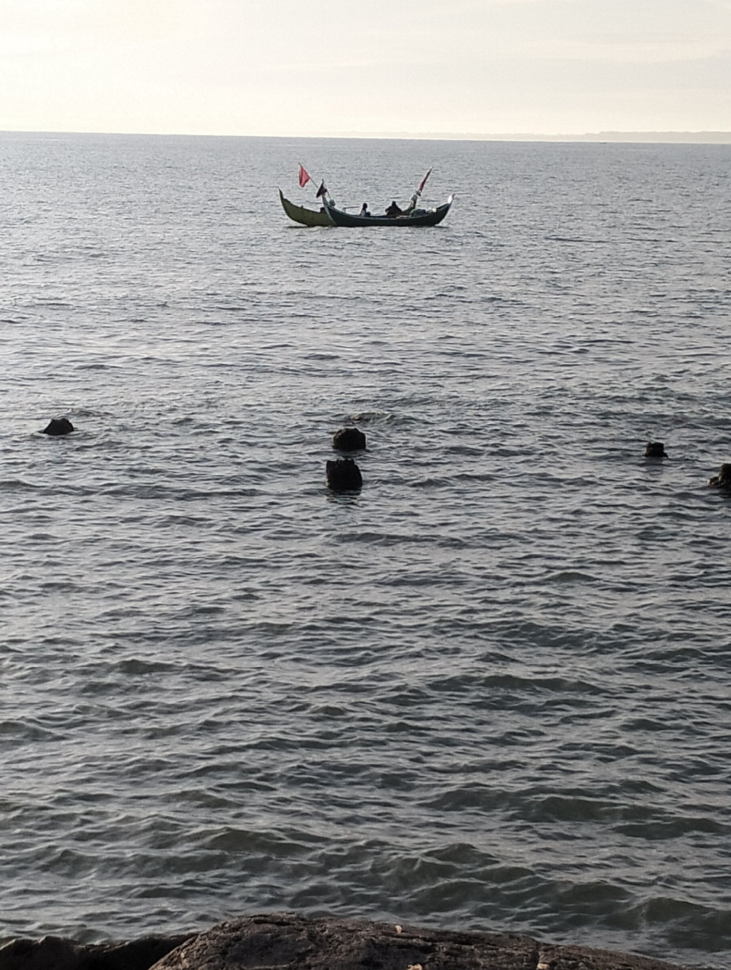 A single, narrow, yellow and dark-colored boat with at least three occupants and a red flag is sailing on a calm, grey body of water. Several dark, angular rock formations are partially submerged in the water at various distances from the camera. The sky is a pale, hazy grey. The scene appears to be a coastal area of Lhokseumawe, Indonesia.