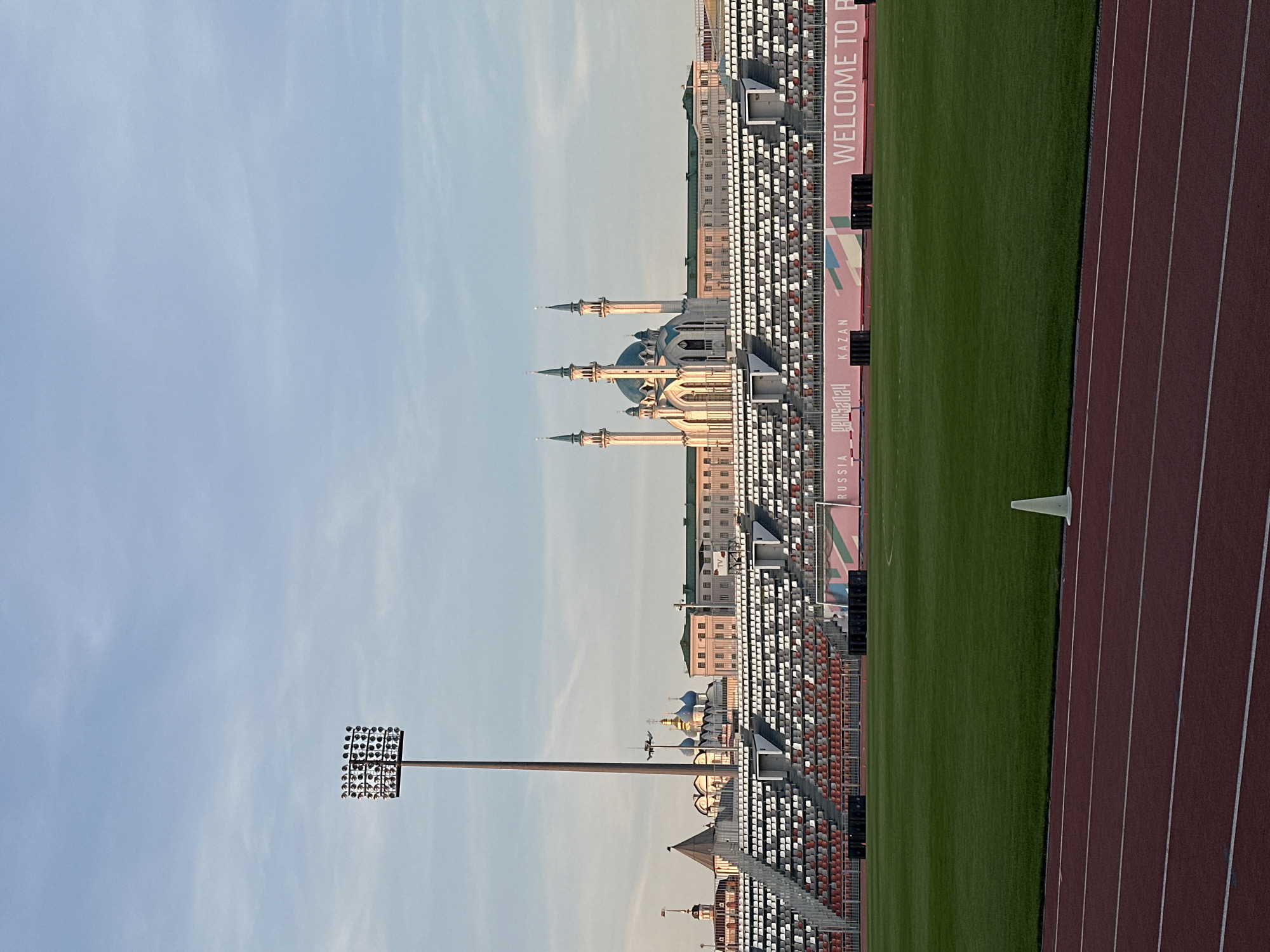The image captures a stadium complex in Kazan, Russia, adjacent to the Kazan Kremlin and Qolşärif Mosque. The stadium features multiple tiers of seating, visible stadium lighting, and a running track bordering a green field. The cityscape backdrop includes various buildings and the distinct skyline of the mosque with its blue domes and minarets. A sign with "WELCOME TO RUSSIA - KAZAN" is displayed along the stadium seating area. The image is taken from a low angle and appears to be oriented vertically.