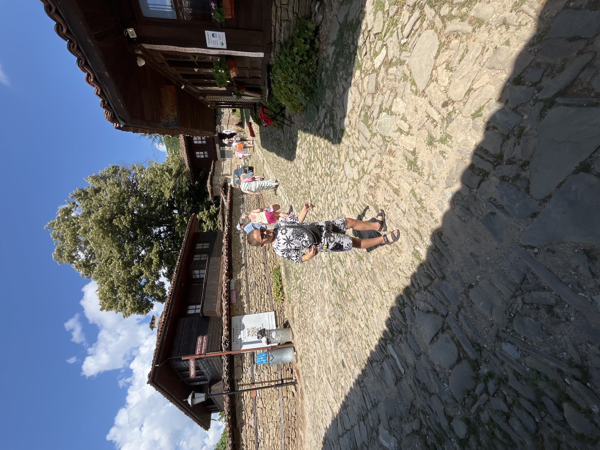 In Zheravna, Bulgaria, a young boy with short, dark hair and sunglasses stands on a cobblestone street, facing the camera. He is wearing a black and white patterned t-shirt and shorts, with black sandals. A black fanny pack is worn across his chest. Behind him, the street is lined with traditional Bulgarian Revival-era houses constructed with stone foundations and dark wooden upper floors, featuring red tile roofs. Several people, seemingly tourists, are visible in the background walking along the street and entering buildings. The sky is blue with scattered white clouds, and a large, leafy tree provides shade. The scene is illuminated by bright sunlight, casting shadows across the cobblestones.