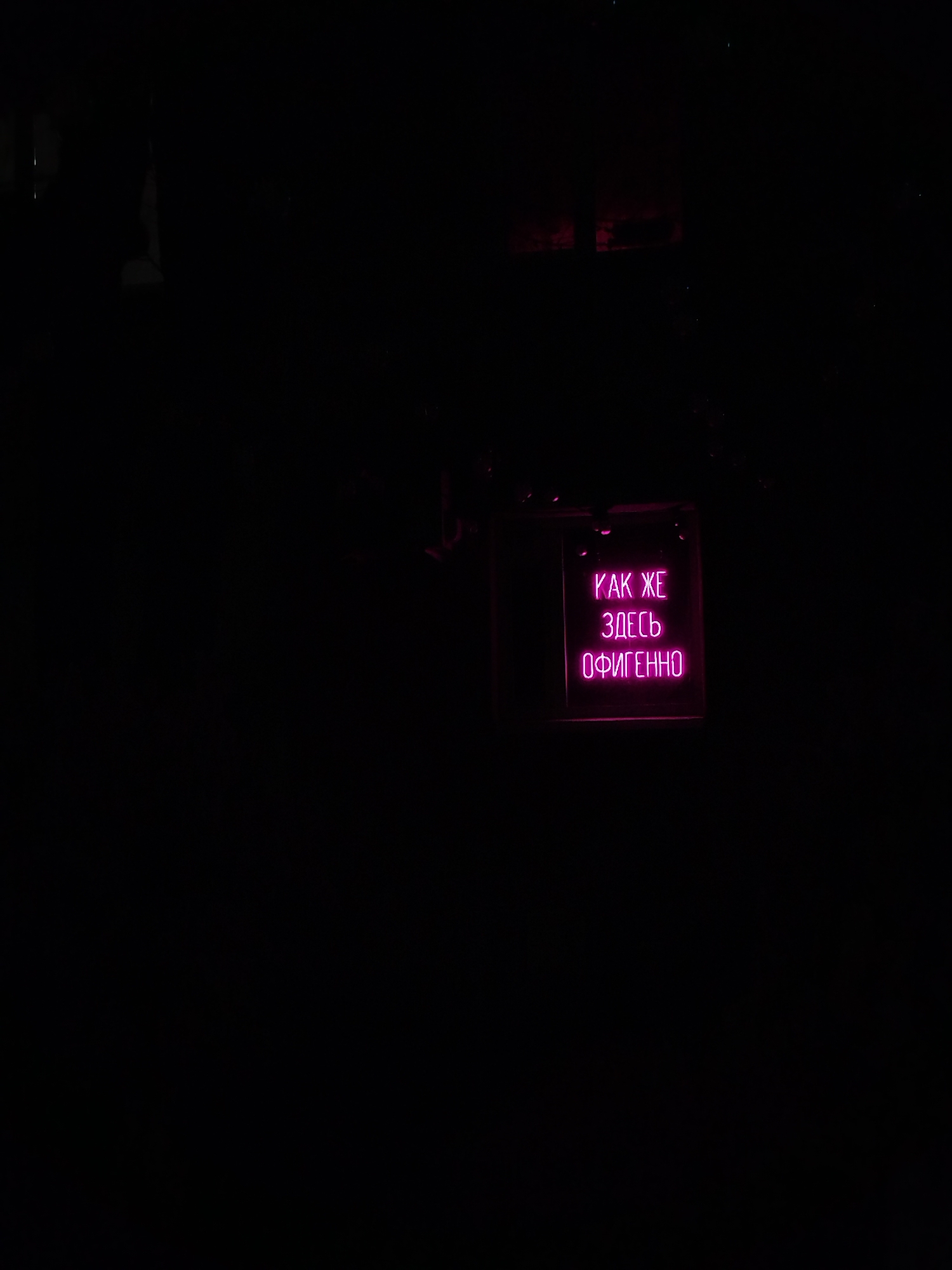 The image displays a dark, interior scene at what appears to be night, dominated by a vibrant pink neon sign. The background is almost entirely black, suggesting very low light conditions or a focus on the illuminated sign. The sign is rectangular, featuring three lines of Cyrillic text: "КАК ЖЕ", "ЗДЕСЬ", and "ОФИГЕННО". This translates to "How awesome it is here" or "It's so f***ing awesome here," conveying a strong positive sentiment.
The neon light casts a soft pink glow, subtly highlighting the immediate area around the sign but leaving the rest of the frame in deep shadow. No people or other distinct objects are visible. The setting evokes the atmosphere of a modern bar, club, or a similar establishment designed for evening entertainment. The vibrant pink color of the neon, combined with the colloquial text, suggests a lively, informal, and perhaps rebellious mood. Given the context of Izhevsk, Russia, the Cyrillic text is a direct cultural cue for the location. The image captures a moment of expressive signage in a nocturnal urban environment.