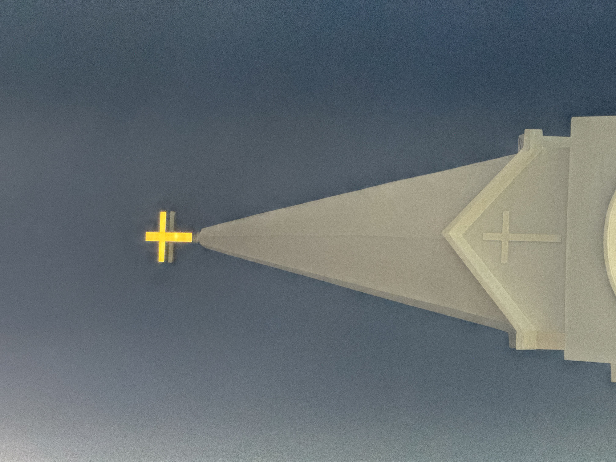 The image captures the spire of a church against a dark blue sky. At the apex of the spire, a bright yellow cross is prominently displayed. Below the yellow cross, further down the spire, a smaller, white cross is visible within a decorative architectural element. The spire itself is pointed and appears to be made of a light-colored material, possibly concrete or stone. The overall scene suggests a religious building situated in an urban environment, though the specific location in Nha Trang, Vietnam, cannot be determined from the visual information provided. There are no people or discernible actions taking place.