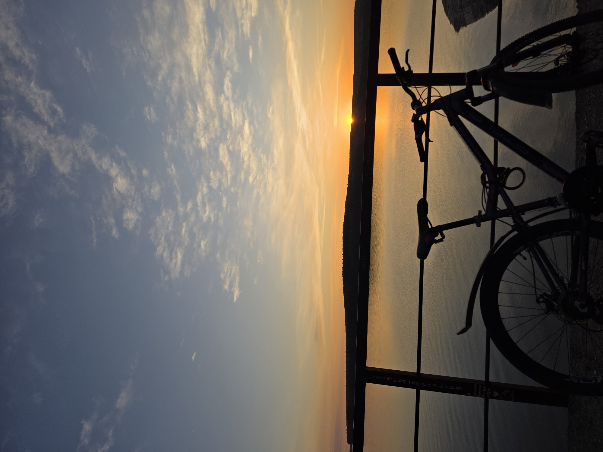 A bicycle is parked against a fence, silhouetted against a sunset. The background consists of a body of water with a strip of land in the distance and a cloudy sky. The scene is located in the city of Chaikovsky, Russia.