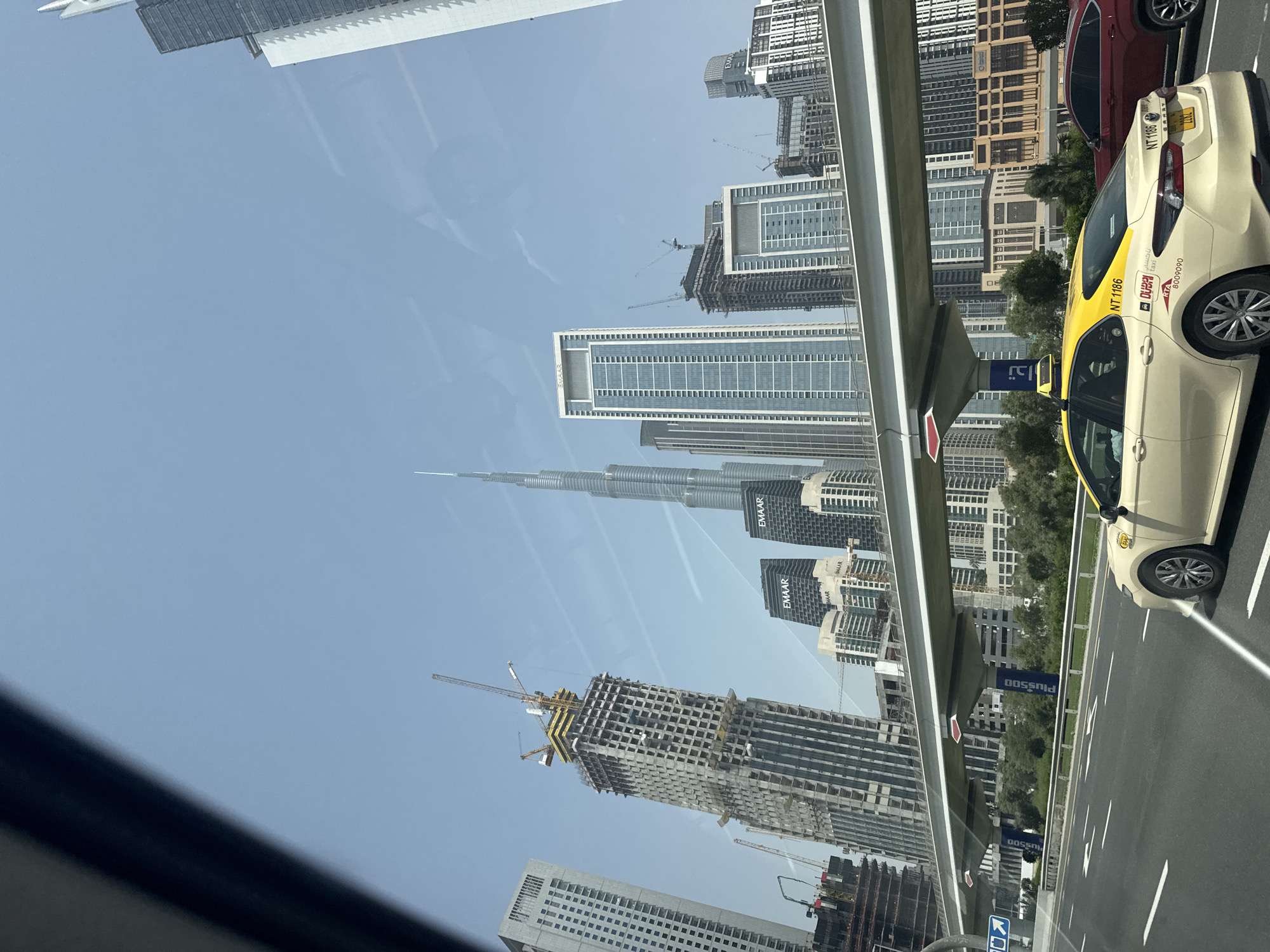 The image, taken from inside a moving vehicle, captures the skyline of Dubai, United Arab Emirates. The most prominent feature is the Burj Khalifa, identifiable by its unique, tapering silhouette. Several high-rise buildings, some still under construction with visible cranes, are also present. In the foreground, a yellow Dubai taxi is positioned on the right side of the road, along with a red car in the lane next to it. The road appears to be a multi-lane highway. A part of the Dubai Metro elevated track is visible. The sky is clear. There are advertisements for Plus500 on the Metro line and the taxi has "NT 1186" displayed.