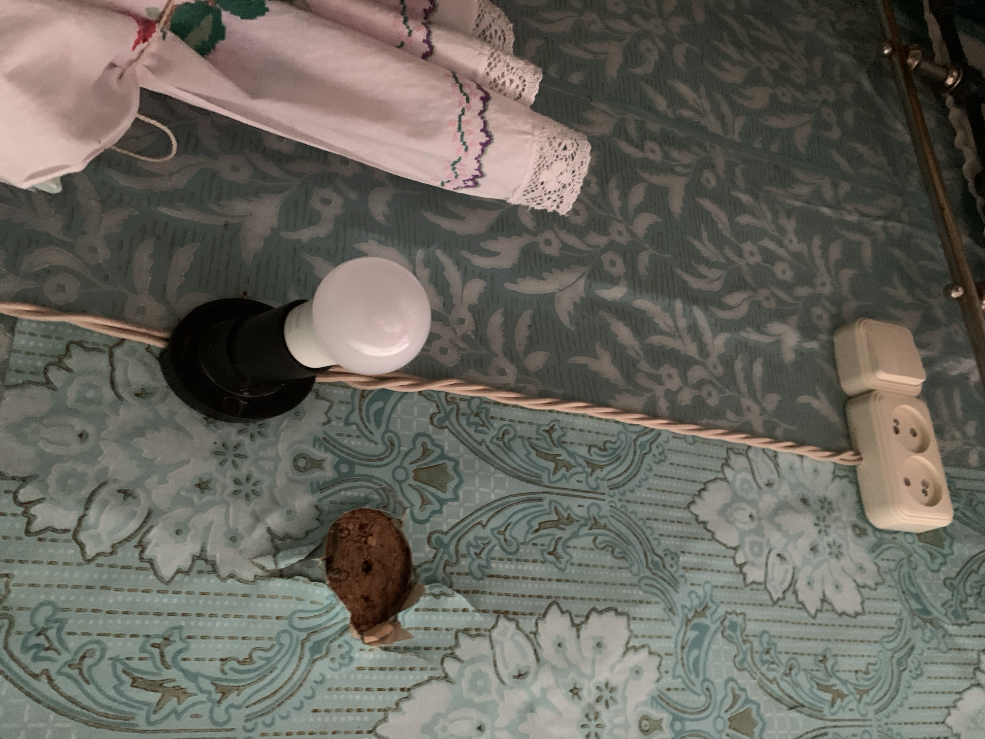 The image depicts a close-up view of a wall covered in patterned wallpaper. A white light bulb is attached to a black fixture, and a braided cord runs vertically along the wall. To the left, a small, brown, circular object with visible holes is attached to the wallpaper. Below the light bulb, there are two white electrical outlets. On the right side of the image, a piece of pink fabric with embroidered detailing and lace trim is hanging. The scene appears to be indoors, possibly in a residential setting, though the specific location in Відзаўскі сельскі Савет, Belarus cannot be determined from the visual information alone.