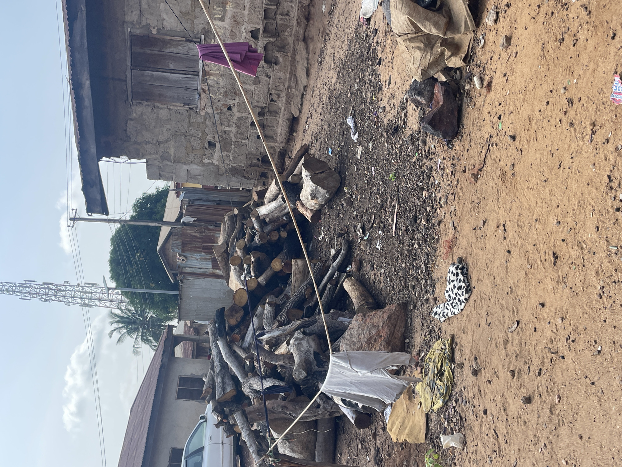 The image depicts an outdoor scene, likely in Lokoja, Nigeria, characterized by a pile of cut logs in the foreground, some of which appear to be split or partially burnt. A white shirt is hanging on a clothesline strung between two points, with a purple garment also hanging from a wire attached to a stone building. To the left, a white car is parked near a building with a corrugated metal roof. In the background, a large metal tower, possibly for telecommunications, rises above palm trees and other vegetation. The ground is composed of reddish-brown earth, gravel, and scattered debris, including pieces of paper and plastic. The architecture visible includes a rough stone building with a wooden shuttered window and what appears to be a more modern building with a metal roof. The sky is partly cloudy.