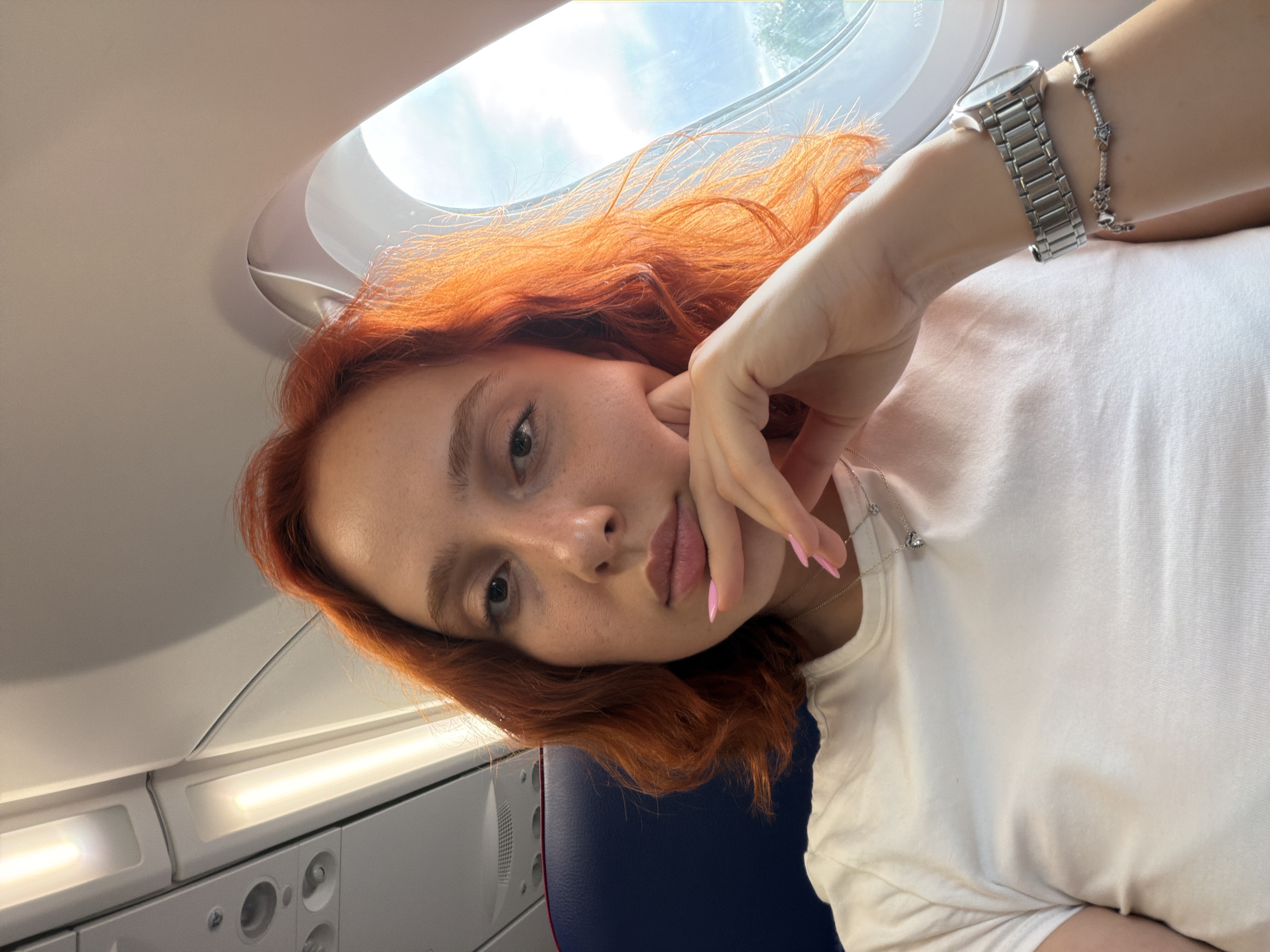 A young woman with vibrant red hair is taking a selfie inside an airplane. She has her head tilted and is resting her chin on her hand, with her fingers gently touching her cheek. Her eyes are open and looking towards the camera. She is wearing a simple white t-shirt and a silver watch on her left wrist, along with a delicate silver necklace. Her fingernails are painted pink.
The setting appears to be a window seat on a commercial airplane. The oval-shaped airplane window is visible in the upper part of the image, revealing a bright sky, likely indicating daytime. The interior of the plane is visible in the background, with a lit panel and a glimpse of a blue seat. The lighting suggests it is daytime, possibly late morning or early afternoon, given the brightness of the sky. The woman's expression is relaxed and contemplative. No visible text is present in the image. The image was taken in Memmingen, Germany.