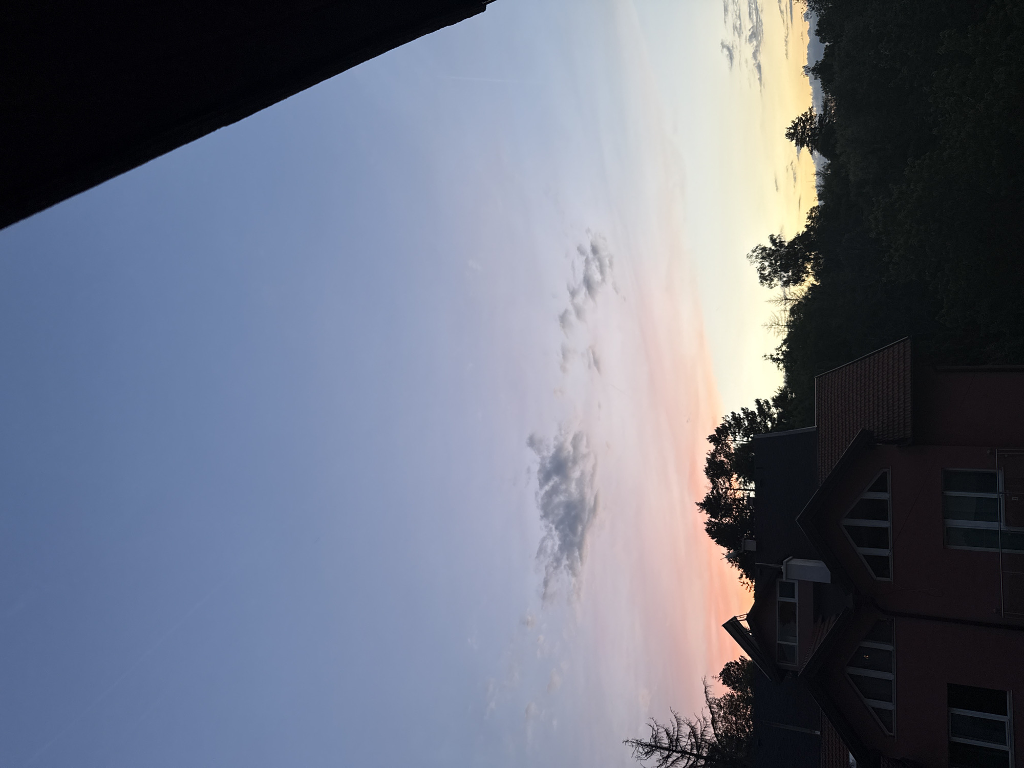 The image depicts a sunset over a residential area in Sofia, Bulgaria. The sky transitions from a deep blue at the top to soft pinks and oranges near the horizon, with scattered clouds. In the foreground, the rooftops of houses are visible, with one house featuring a prominent red roof and triangular attic windows. Trees and the silhouettes of more houses and structures are nestled within a dark, verdant landscape that occupies the lower portion of the image. The scene is serene, capturing the quiet transition from day to night.