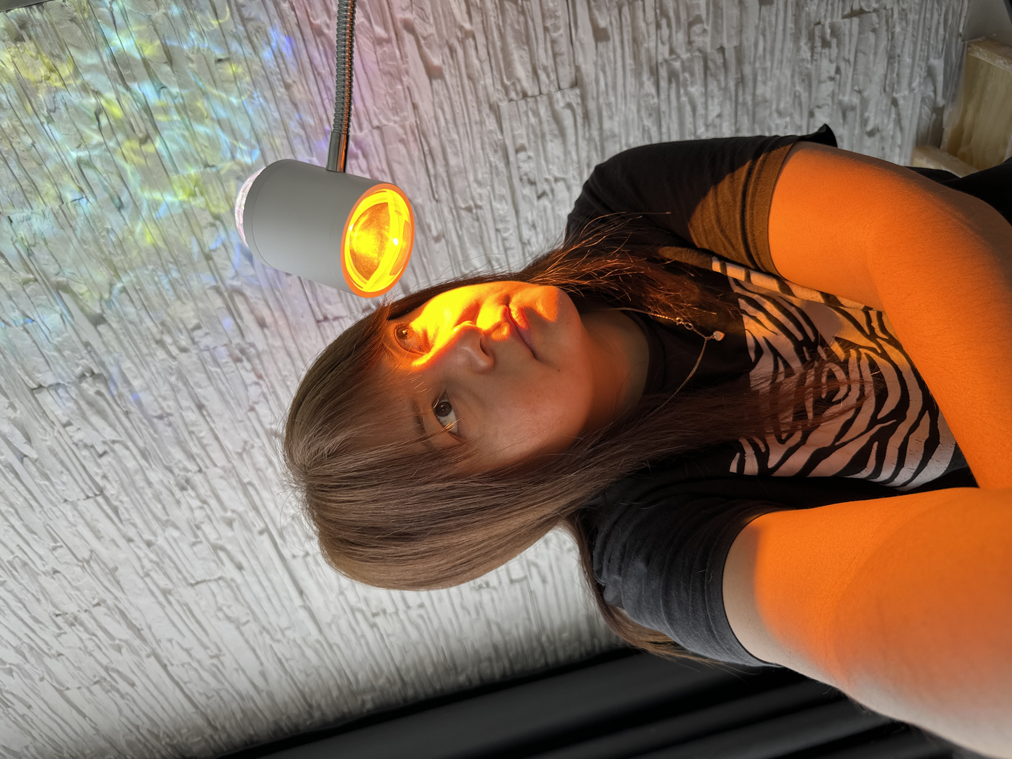 A rotated, close-up shot shows a person illuminated by a warm light emanating from a small, white cylindrical lamp attached to a flexible metal arm. The light source is positioned above and slightly to the left of the person's face, casting a strong orange glow. The person has light brown hair with bangs, fair skin, and is wearing a dark shirt with a black and white zebra-stripe pattern on the shoulder and sleeve. The background consists of a wall with a textured, light-colored surface, part of which is illuminated with abstract patterns of blue, yellow, and white light. The overall composition suggests an indoor setting. The city and specific location are unknown.