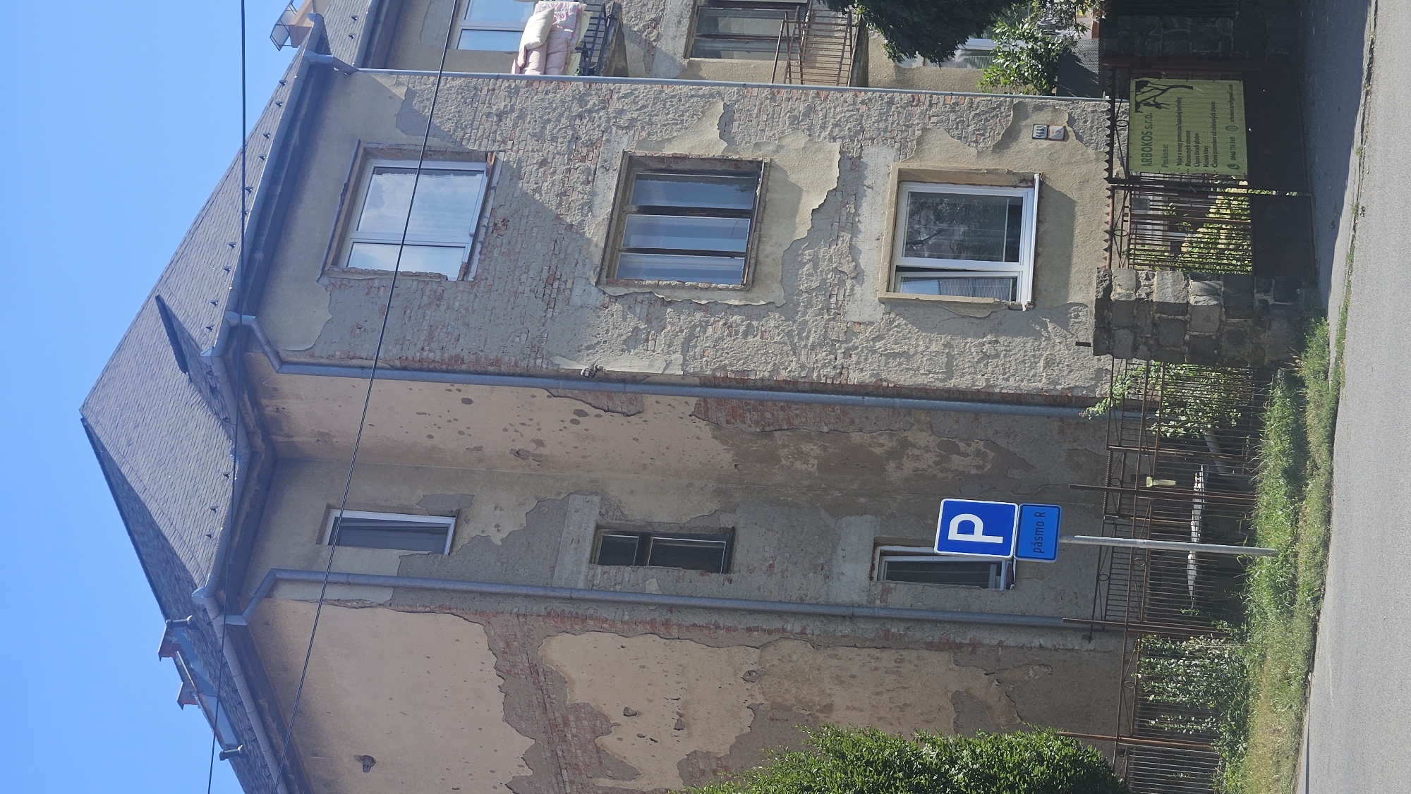 The image shows a multi-story building in Prešov, Slovakia, with a weathered facade. The stucco is peeling, revealing brickwork underneath in several areas. The building has a dark gray, gabled roof with visible gutters and downspouts. There are multiple windows of various sizes and styles on the building, some appearing older and others newer.
In the foreground, there is a blue parking sign with a white "P" and the text "pásmo R" below it. A metal fence runs along the property line, separating the building from a grassy area and a paved road. To the right of the fence, there is a gate with a yellow sign that reads "ARBAKOS S.R.O." and lists services such as "Výsadba a nová profesionalizovaná údržba," "Predaj stromov a kríkov," and "Kosenie trávy." There are also contact phone numbers and an email address on the sign.
A lush green tree is visible on the left side of the image. The sky is clear and blue, indicating a sunny day. The overall impression is of an older urban building with some signs of wear and tear, situated in a residential or commercial area of Prešov.