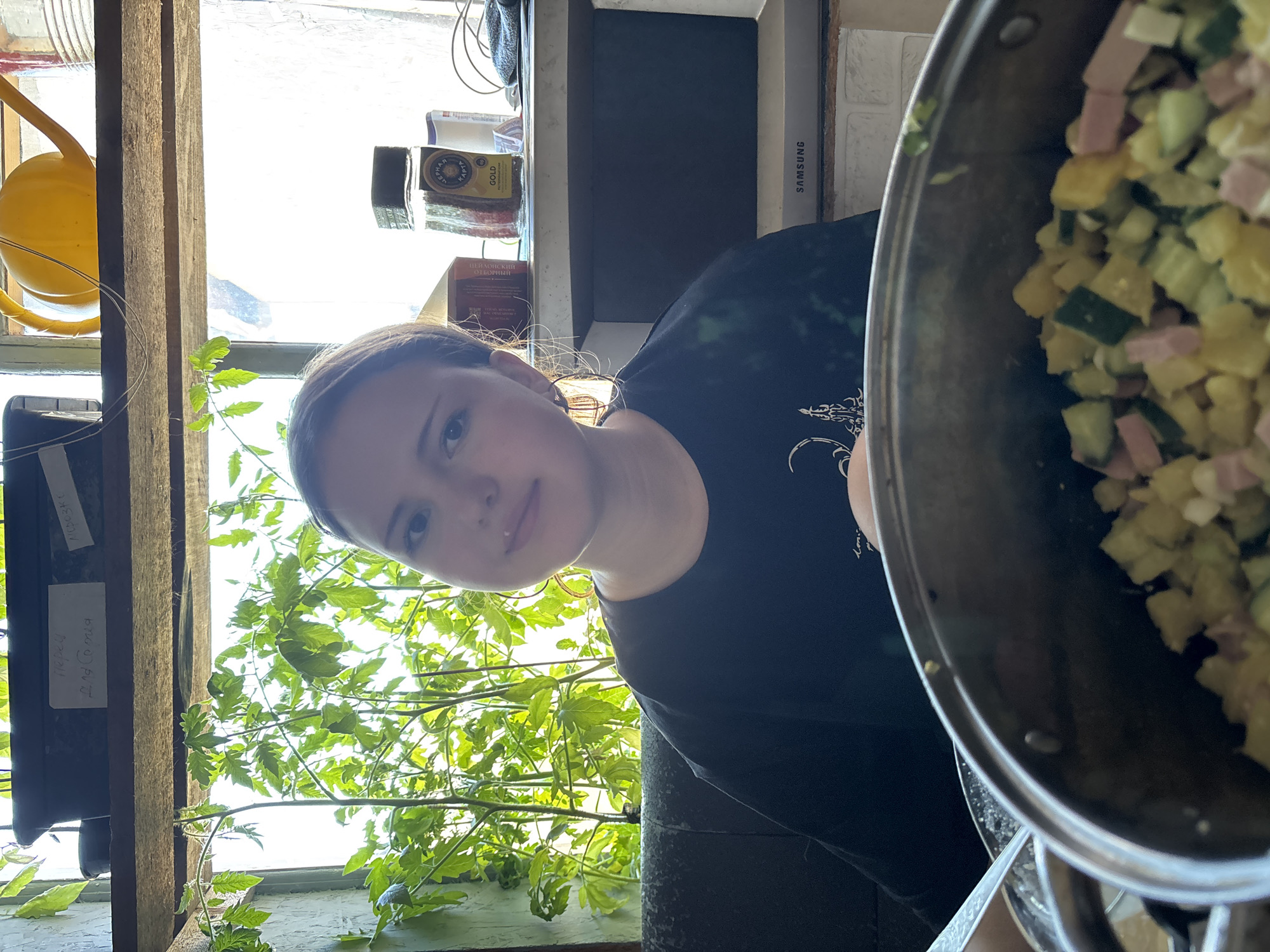 A young woman with fair skin and light brown hair wearing a black t-shirt is pictured holding a large metal pot containing a mixture of diced zucchini and ham. The scene is brightly lit by natural light streaming through a window behind her, which reveals lush green foliage. Visible in the background, behind the woman, are items such as a Samsung monitor, jars of spices, and a yellow watering can. Labels in Russian are visible on a planter. The location is likely within a domestic setting. The scene appears to be in Фоки, Russia.
