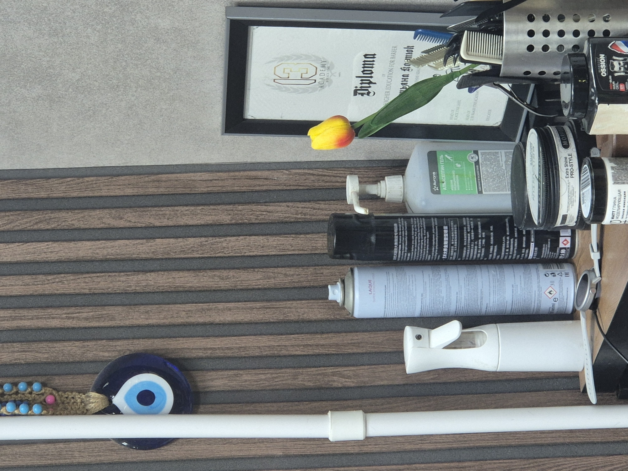 The image shows a shelf with various items, likely in a barbershop or salon, in Kokshetau, Kazakhstan. The background features a vertically slatted wooden wall. On the left, a white pole is visible, with a blue and white evil eye charm hanging from it. Next to the pole is a white spray bottle.
Moving to the right, there are several aerosol cans. One is light blue with "LAQUE" branding. Another is black with Korean and English text. A clear pump bottle with a green label reading "AURORA АЛСПЕПТИН ГЕЛЬ" is also present.
Further to the right, a diploma framed in black is displayed. The diploma has "ACADEMY BY BLACK STAR" and "Diploma" text visible, along with other Cyrillic script. A single yellow and red tulip is placed in front of the diploma.
In the foreground, there are several hair styling products in tubs. One tub is labeled "MOREFOSE EXTRA SHINE PRO-STYLE". Another smaller tub is labeled "МАТТ ГЛИНА МОДЕЛЛИРУЮЩАЯ". To the right, there is a metal cup filled with various combs and scissors. A black bottle with a label featuring a barber's silhouette is partially visible at the bottom right. A smartwatch is also visible on the shelf in the foreground. There are no people in the image.