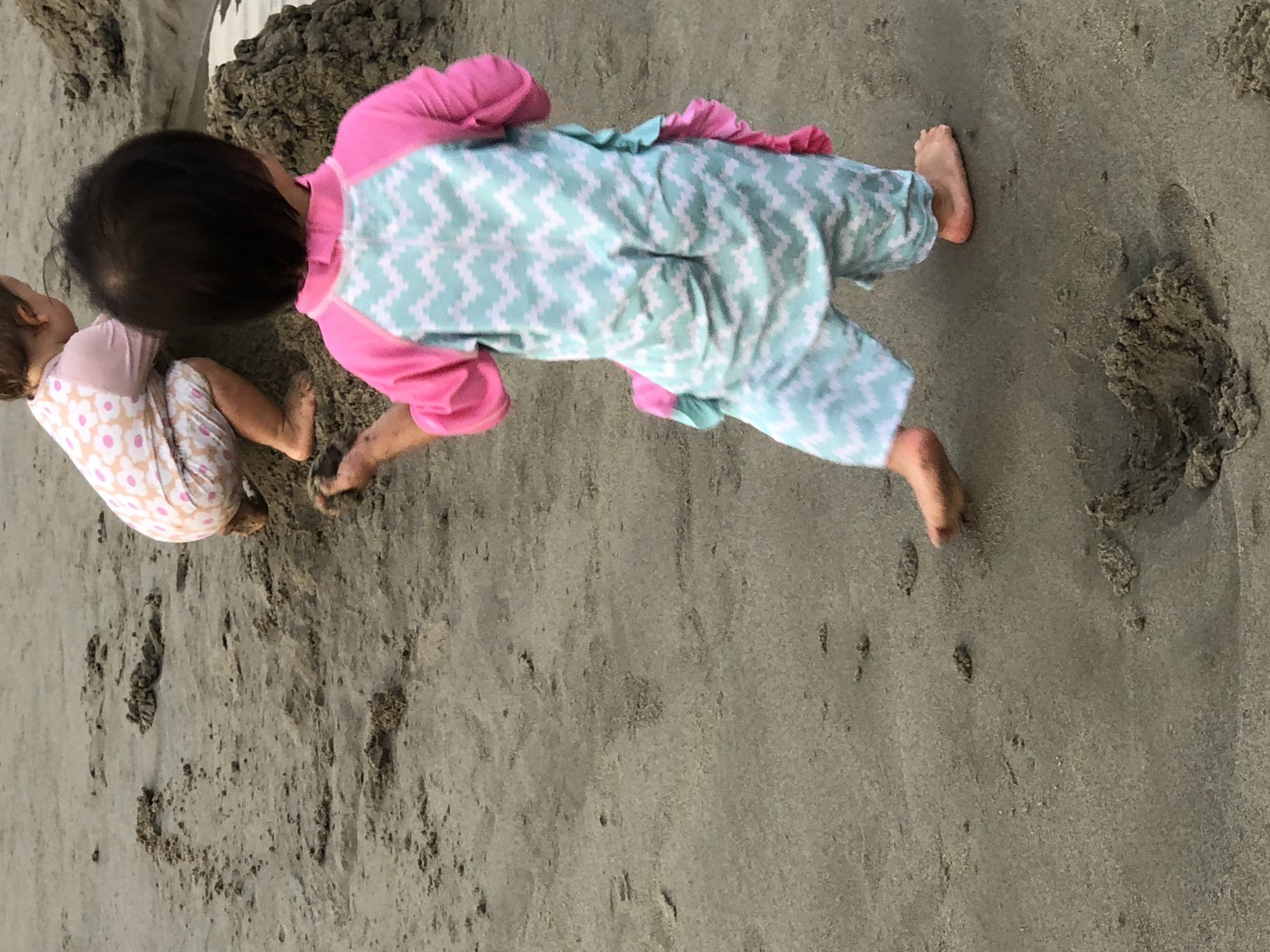 Two toddlers are playing in the sand on a beach. The toddler in the foreground is wearing a pink and aqua chevron print rash guard and swim trunks. The toddler in the background is wearing a pink and white floral print rash guard and swim trunks. They are both digging in the sand and appear to be enjoying themselves. The sand is wet and there are footprints in the sand. The image was taken in Da Nang, Vietnam.