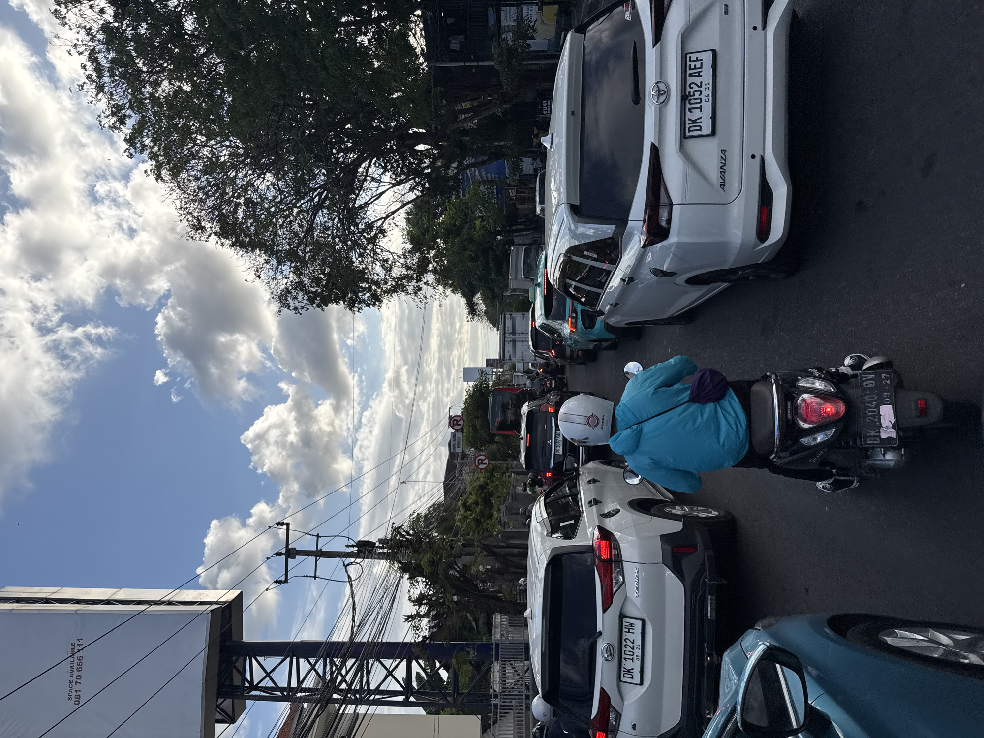 The image depicts a street scene in Denpasar, Indonesia, characterized by moderate traffic congestion. In the foreground, a person on a black scooter wearing a turquoise jacket and a white helmet is navigating through traffic. Behind the scooter, a white Toyota Terios SUV with license plate DK 1022 HW is visible. To the right, another white Toyota Avanza SUV with license plate DK 1052 AEF is also in traffic. The road is asphalt, and the sky above is partly cloudy. A large billboard with "SPACE AVAILABLE" and a phone number is present on the left side of the image, along with numerous power lines strung between poles. In the background, trees line the street and other vehicles, including a bus, are part of the traffic flow. The overall atmosphere suggests a typical urban commute in Denpasar.
