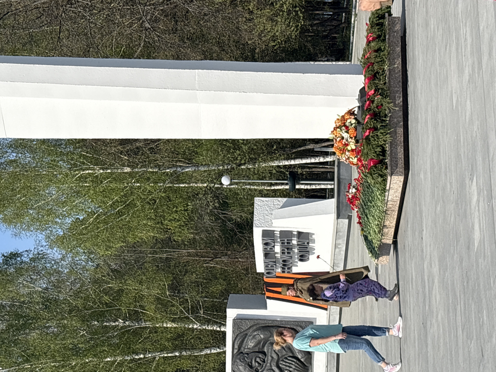 The image shows a monument in the city of Chaikovsky, Russia. The monument consists of a tall white rectangular block and adjacent gray stone structures with inscriptions in Cyrillic and a bas-relief. Wreaths and greenery adorn the monument. Two people stand in the foreground. One is an adult female with blond hair in a ponytail, wearing a turquoise t-shirt, blue jeans, and white sneakers. The second person is a child in a purple patterned outfit with a brown jacket and a black and orange striped ribbon. They appear to be laying a single red flower at the base of the monument. Trees are visible in the background. The sky is clear with blue and white colors.