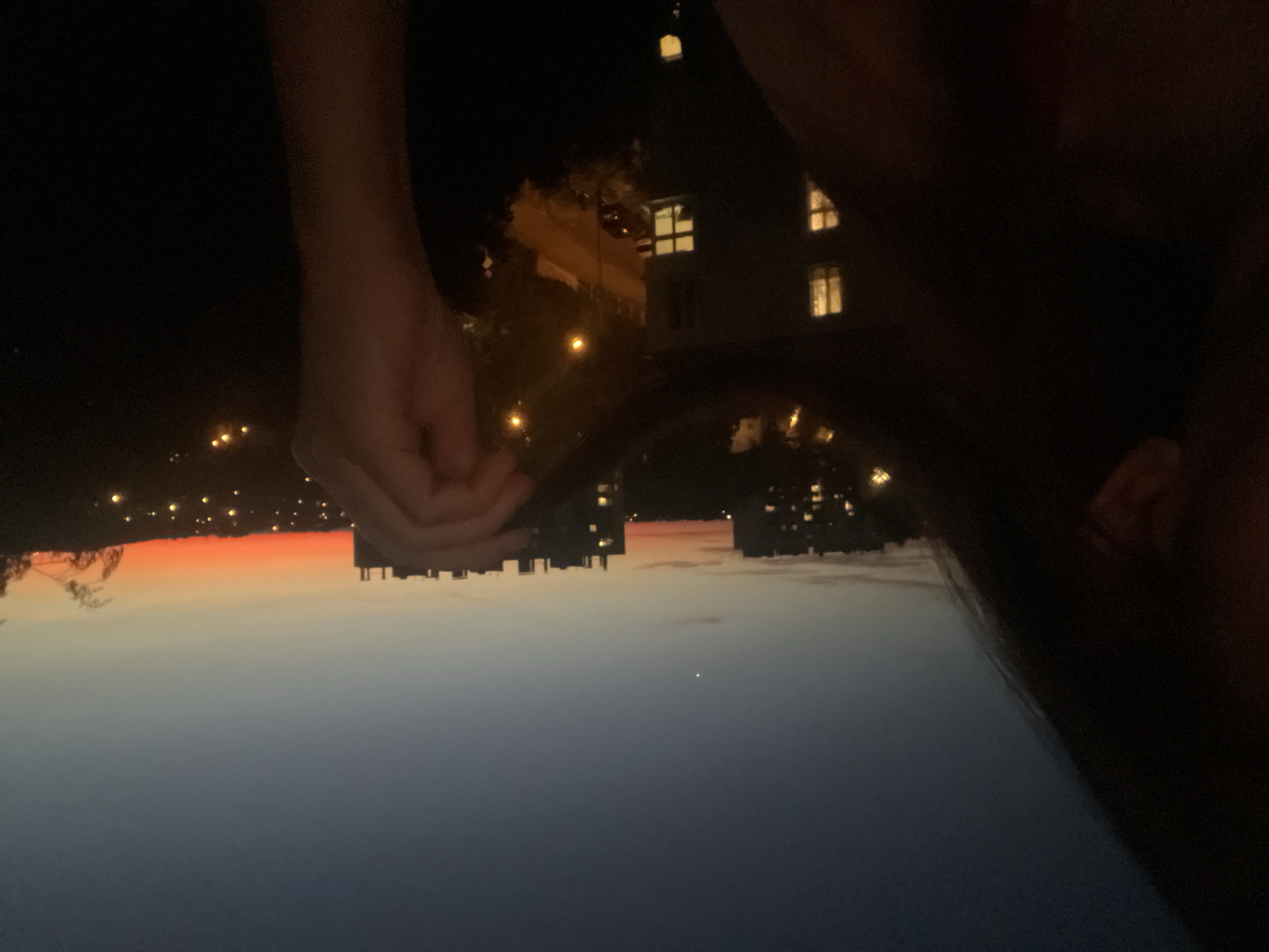 The image is a nighttime shot with a focus on a person's arm and hand, partially obscuring a cityscape and a twilight sky. The composition is inverted, with the sky appearing at the bottom and the cityscape at the top.
The person's arm and hand are prominently featured in the upper left quadrant of the image, appearing pale in the low light. The hand is open, fingers slightly curled.
The background consists of buildings with illuminated windows, suggesting an urban setting. Streetlights and other small lights are visible throughout the scene, dotting the landscape like scattered stars. The sky displays a gradient of colors, transitioning from a deep blue at the bottom to a bright orange band near the horizon, characteristic of twilight or dusk. A single bright point, possibly a star or planet, is visible in the blue expanse of the sky.
The scene evokes a sense of quiet contemplation, with the darkness of night contrasting with the artificial lights of the city and the fading light of the sky. The presence of the person's arm and hand suggests a personal perspective, as if the viewer is looking over their shoulder or through their arm. The overall mood is somewhat mysterious and peaceful, with the inverted orientation adding a slightly disorienting yet intriguing quality. No visible text is present in the image.