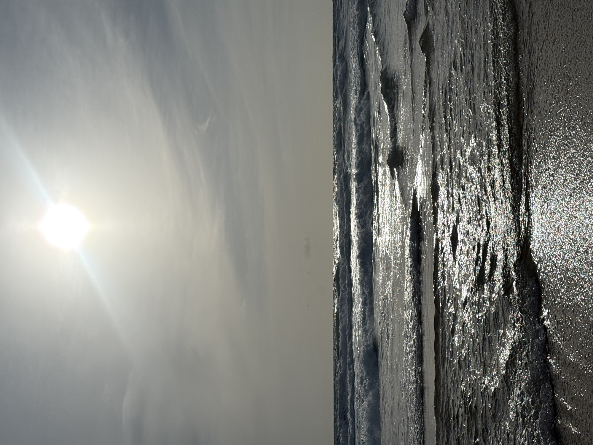 The image shows a beach scene at Karon, Thailand. The sun is high in the sky, partially obscured by clouds, and its rays create a bright glare. Gentle waves roll in towards a sandy shore, their surfaces reflecting the sunlight with a shimmering effect. The water appears dark and somewhat opaque, with white foam cresting on the waves. The sky is a pale gray, with wispy clouds. There are no people or man-made objects visible in the frame, suggesting an undeveloped stretch of coastline.