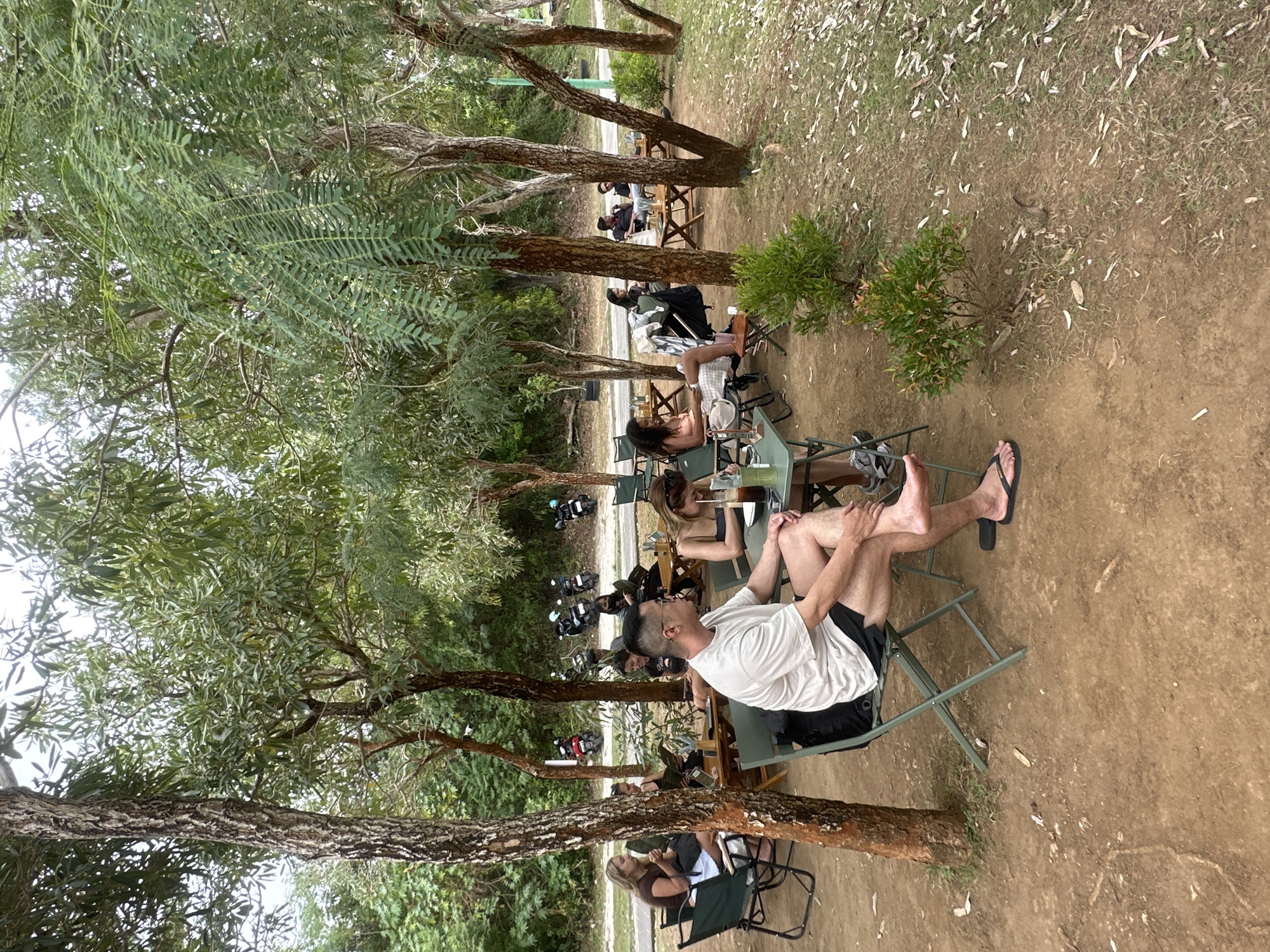 A group of people are seated at outdoor tables under trees in Kuta Selatan, Indonesia. The scene appears to be a cafe or restaurant with dirt ground and sparse grass. In the foreground, a man in a white t-shirt and black shorts sits in a green chair, facing right, with his legs crossed. Behind him, other patrons are seated at tables, some looking at menus, others engaged in conversation. Several motorcycles are parked in the background. The environment is lush with trees and foliage, suggesting a tropical setting.