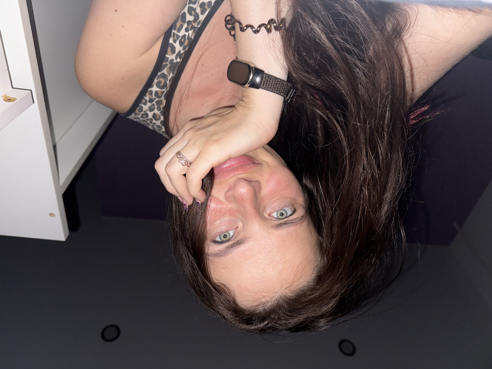 A woman with long, dark brown hair and green eyes poses for a selfie in a dimly lit room. She is resting her chin on her hand, wearing a leopard print tank top. A ring with a heart design is on her right ring finger. A fitness tracker is on her left wrist, and a coiled bracelet is below it. The background is dark, with a white shelf or cabinet visible on the right side of the frame. There is also a circular light fixture visible on the ceiling.