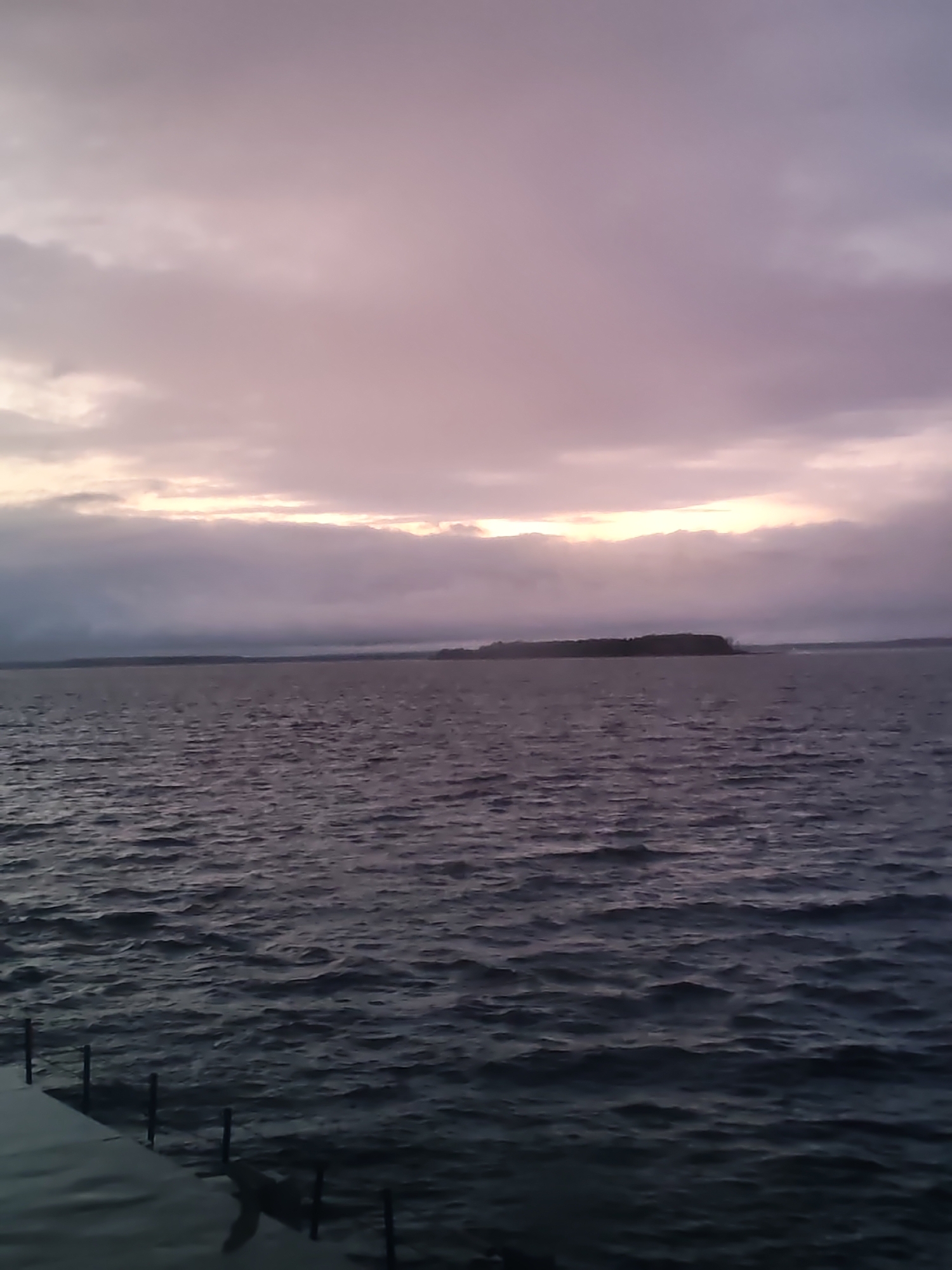 The image captures a serene landscape scene dominated by a vast body of water under a cloudy sky, likely at dawn or dusk. The water, which occupies the lower two-thirds of the frame, is dark and exhibits numerous ripples, indicating slight movement or a gentle breeze.
Across the distant horizon, a low, dark landmass, possibly an island or a far shore, is visible, appearing as a long, flat silhouette against the sky. The sky itself is heavily overcast with layers of clouds, predominantly in shades of soft purple, grey, and hints of pale pink. A distinct horizontal band of diffused light, characterized by pale yellow and faint orange hues, breaks through the clouds near the horizon, suggesting the position of the sun during a sunrise or sunset. This light illuminates the undersides of the clouds, creating a soft glow.
In the bottom-left corner of the image, a dark, angled structure, consistent with a dock or pier, extends into the water. Several vertical posts or pilings are visible along its edge. No people or animals are present in the scene. The overall atmosphere is peaceful and contemplative. No text is visible.