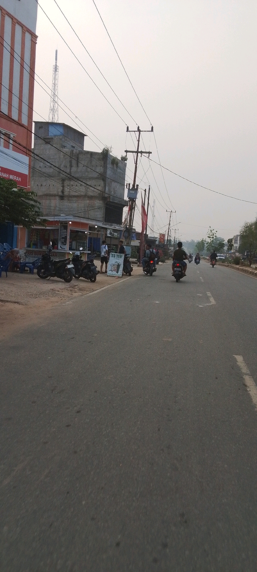 The image captures a street scene in Lenggadai Hulu, Indonesia. A paved road dominates the foreground, with a slightly hazy, overcast sky filling the upper portion of the frame. On the left side of the road, a multi-story building with an orange and white facade stands next to a rougher, concrete building with visible electrical conduits. In front of these buildings, several motorcycles are parked along the roadside. A small food stall with a colorful awning is visible, and a sign advertising "ES TEH" (iced tea) stands near the entrance.
Several people are present. Near the food stall, three individuals stand and interact. One person is seated on a motorcycle, while another is walking away from the stall. Further down the road, several other individuals are riding motorcycles, moving away from the camera. One motorcyclist is in the middle of the road, while others are further in the distance. Electrical poles with numerous wires are scattered throughout the scene, indicating the presence of infrastructure. The environment appears to be a commercial or semi-urban area, with businesses and residential structures lining the street. The overall impression is that of a typical day in a developing urban area.