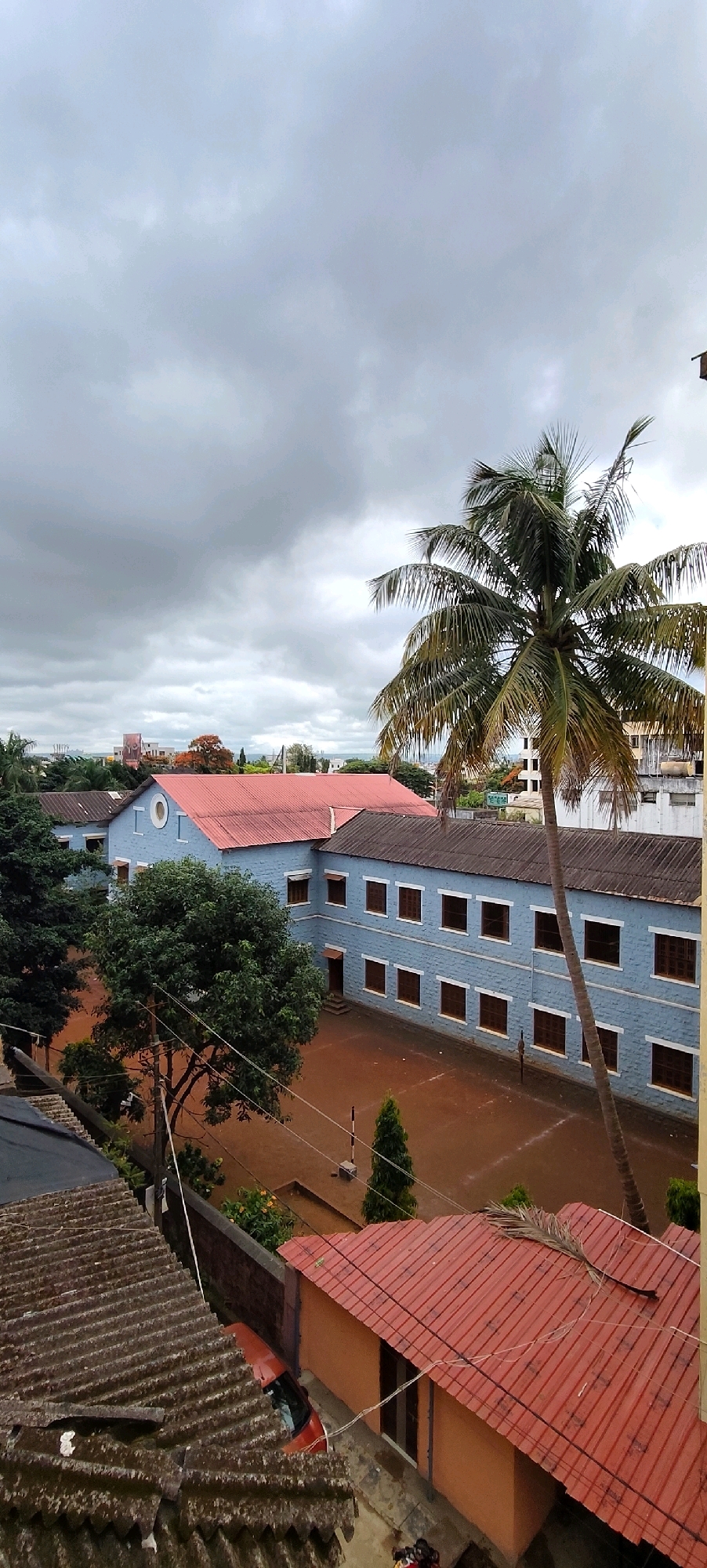 This image captures an overcast day in Belagavi, India, likely from an elevated viewpoint. In the foreground, a corrugated metal roof dominates the bottom left of the frame, showing signs of age and weathering. Adjacent to it, a small orange building with a red tiled roof is visible. A portion of an orange car is parked beside this building.
The mid-ground features a large, blue building with a red-tiled roof, suggesting it might be a school or institution. This building has multiple windows with dark frames. A courtyard in front of this building appears to be muddy. A tall palm tree stands prominently to the right of the blue building, its fronds extending towards the sky. Several green trees are also present, including a dense tree to the left of the courtyard.
In the background, more buildings and trees are visible, hinting at a more developed urban or suburban area. The sky is heavily clouded, with shades of grey indicating a potential for rain or a generally gloomy day. The overall scene depicts a residential or institutional area with characteristic Indian architecture and vegetation.