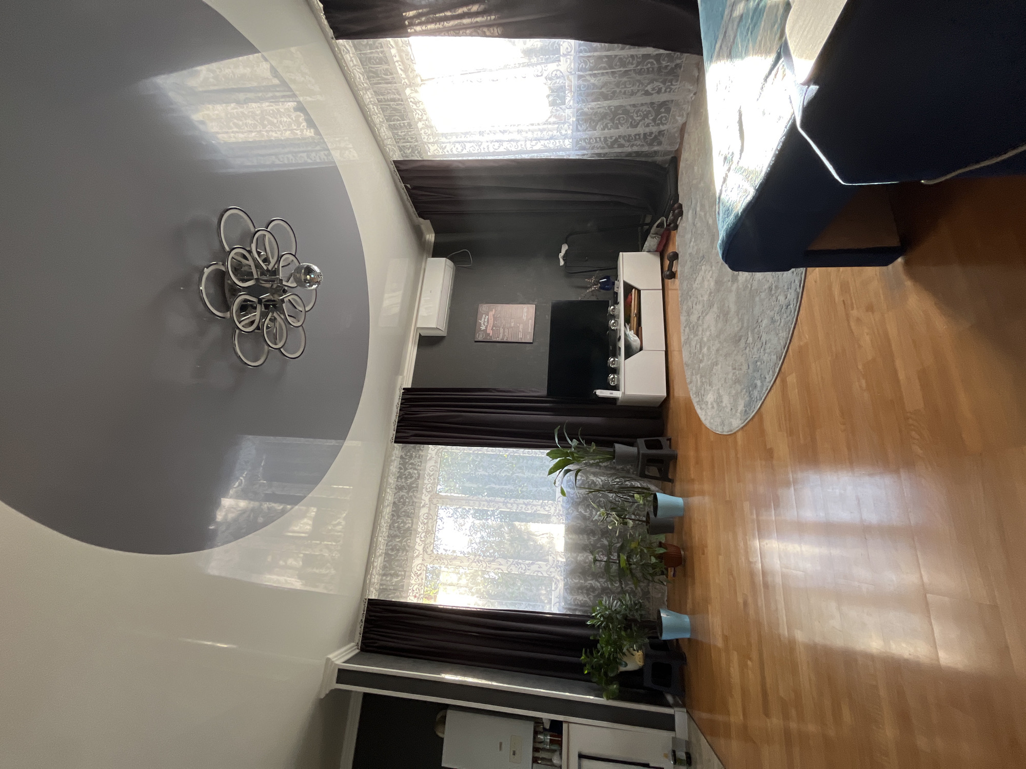The image shows a living room in an apartment in Saratov, Russia. The room has a glossy, multi-level ceiling with a dark grey circular section in the center, from which a modern chandelier hangs. Sunlight streams in through a large window covered by sheer lace curtains and dark brown drapes. On the left, there's a white appliance mounted on a dark grey wall, possibly a water heater. A row of potted plants sits on the light brown wooden floor in front of the window. In the center of the room, a white TV stand holds a television, and a round grey rug is placed in front of it. To the right, a dark blue couch is partially visible, illuminated by the sunlight. The room appears to be tidy and well-lit.