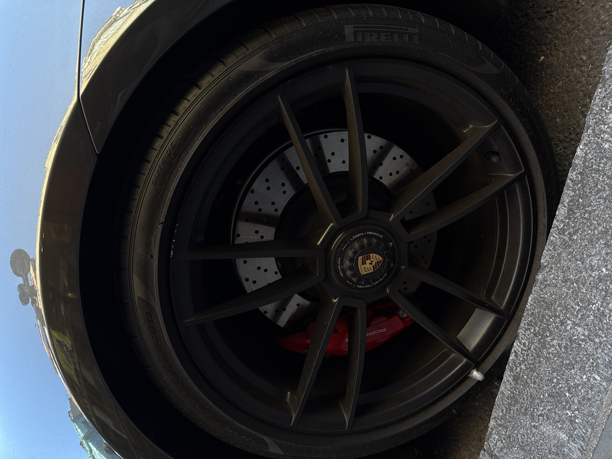 An angled, close-up shot captures the front left wheel of a high-performance car, likely a Porsche, parked on a street in London, United Kingdom. The image is taken from a low perspective, looking upwards and across the wheel.
The prominent subject is a large, matte black, multi-spoke alloy wheel with a center-lock mechanism, rather than traditional lug nuts. The center cap features the distinctive gold and black Porsche crest. Around the center cap, text is visible, including "LÖSEN / REMOVE" (German/English for loosen) and "ANZEIHEN / TIGHTEN" (German/English for tighten), along with "CENTERLOCK" and directional arrows.
Behind the spokes, a large, silver, perforated brake rotor is visible, indicating a performance braking system. The brake caliper is painted bright red and clearly displays the "PORSCHE" branding in white text.
The tire is a Pirelli P Zero, with the brand name "PIRELLI" and "P ZERO" embossed on the sidewall. Other visible text on the tire includes "OUTSIDE," "RADIAL TUBELESS," "XL," and the size "285/30 R20." The tire shows some signs of use, including a light coating of dust.
The car's dark-colored fender arch is visible above the tire, reflecting a bright, clear blue sky, suggesting it is daytime with good weather. To the right of the wheel, a grey, textured curb or sidewalk edge is visible, indicating the car is parked alongside a road. A small, white, fluffy piece of debris is caught between the tire and the curb. Reflections on the car's bodywork also show hints of buildings or streetlights in the background.