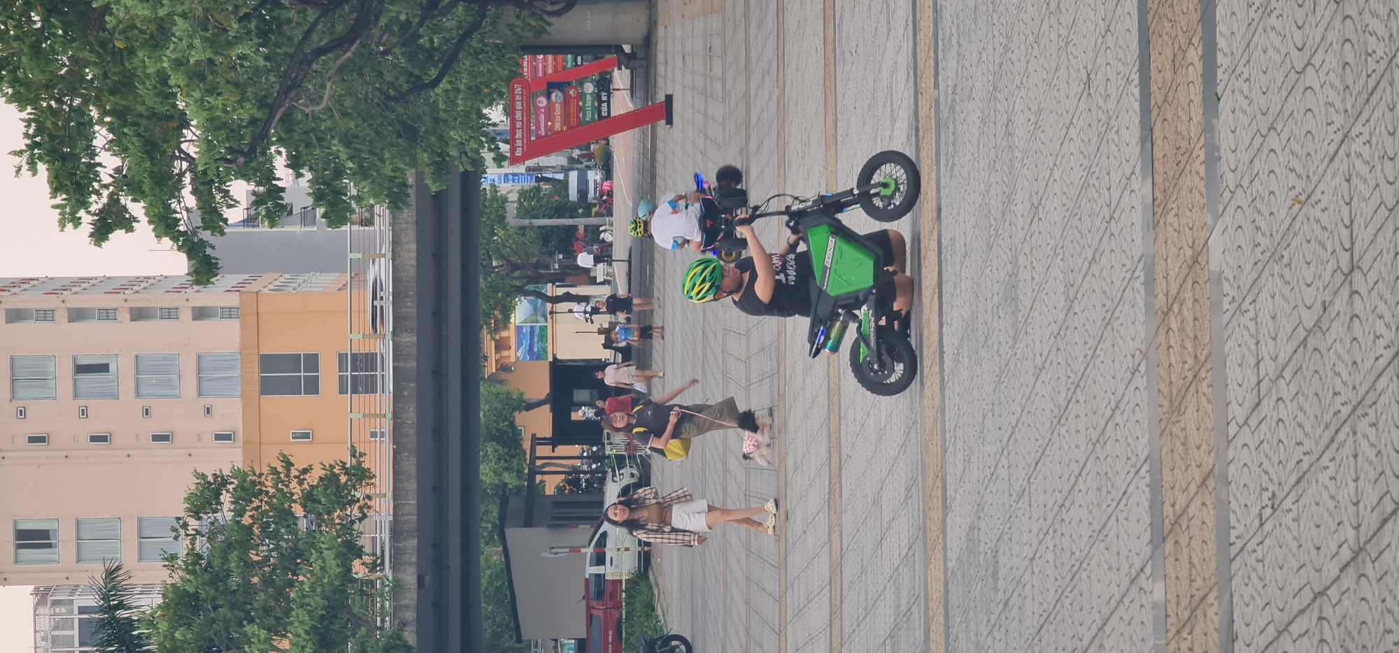 The image depicts a street scene in Đà Nẵng, Vietnam, with a variety of people and objects. A green electric scooter is prominently featured, with a person wearing a black shirt and a green helmet standing behind it. Another person in a white shirt and helmet is riding an electric scooter in the background. Pedestrians are strolling along the tiled pathway, including a woman wearing a white top and shorts, another individual walking a small dog, and additional people in casual attire. In the background, there are buildings, trees, an elevated road structure, and red signage.