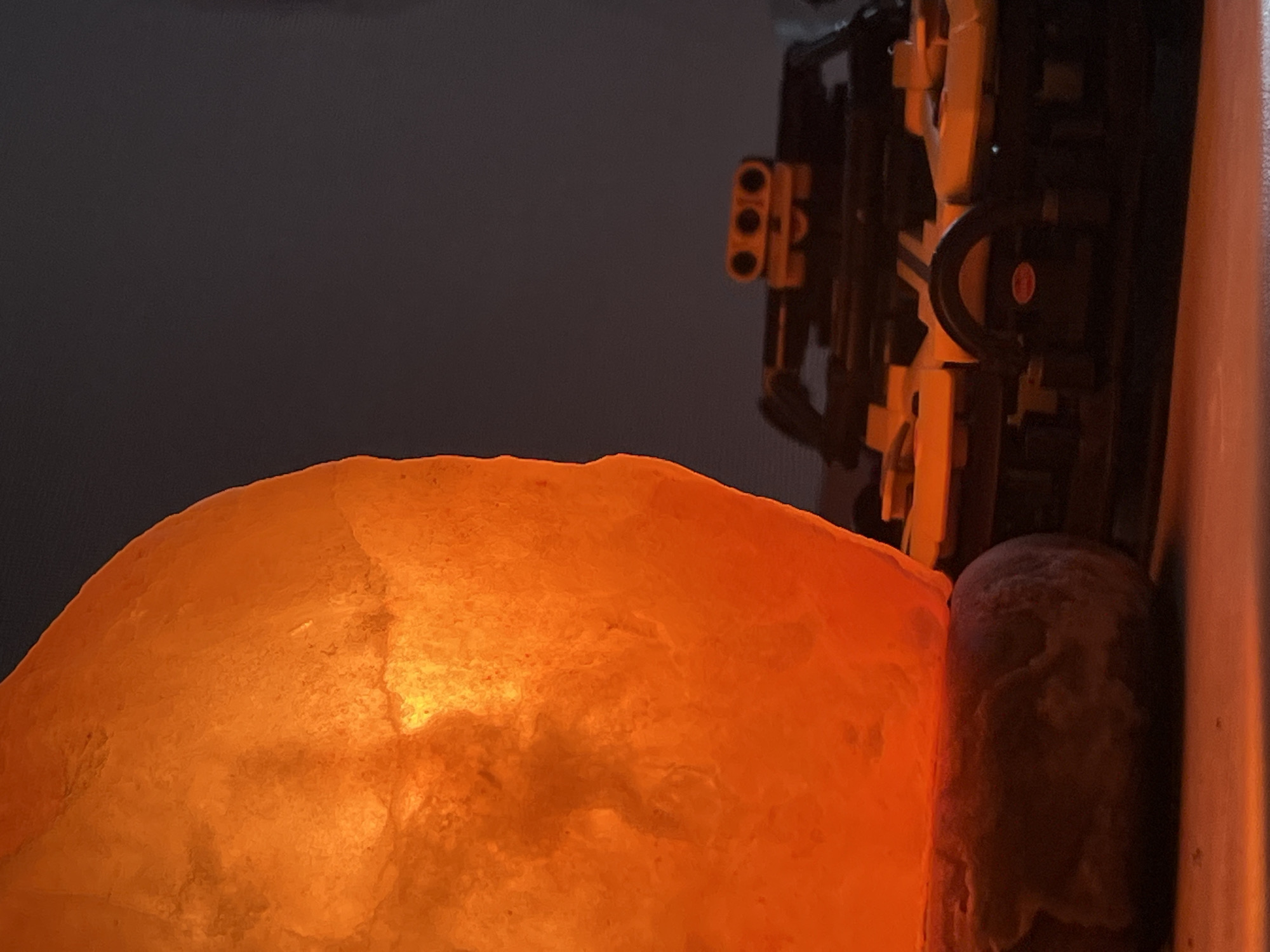 A close-up, rotated shot displays a dimly lit scene featuring a glowing orange object and parts of a mechanical structure. The dominant feature on the left is a large, irregularly shaped object emitting a warm, bright orange light, characteristic of a Himalayan salt lamp. Its surface appears crystalline and textured.
To the right, partially visible, is a complex mechanical assembly constructed from yellow and black components, resembling a building block system like LEGO Technic. Several interlocking pieces, gears, and pins are discernible. In the center-right, a black, ring-like part features a small, red circular sticker with faint white markings, too indistinct to read. Further right, a dark, cylindrical object with a textured or frosted surface rests between the glowing lamp and the mechanical structure. The background is a uniform dark gray, suggesting the image was taken in a low-light environment, possibly at night or in a dimly lit room, with the glowing object serving as a primary light source. No discernible activity is taking place; the image captures a still arrangement of objects.