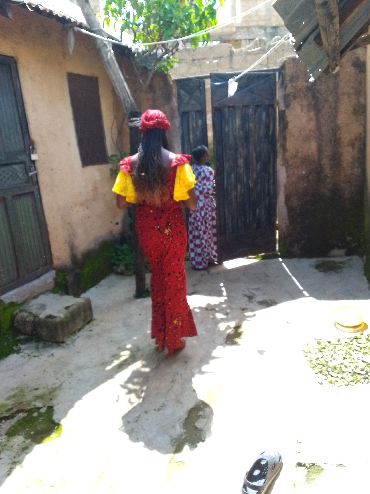 Two women are standing in an outdoor courtyard. The woman in the foreground is facing away from the camera, wearing a red headscarf, a red and yellow dress with yellow ruffled sleeves, and a long, patterned red skirt. The second woman is further back, facing left, wearing a white and red patterned dress. They are standing on a concrete surface with scattered debris and some patches of green moss. Behind them are two large wooden doors, and to the left, a tan-colored building with a dark green door and a window. To the right of the doors, the wall appears to be made of rough stone blocks. Overhead, a corrugated metal roof is visible, supported by wooden beams. Sunlight casts shadows on the ground. In the bottom right corner of the image, a pair of dark and white patterned shoes is visible on the ground. The location is identified as Unknown, Unknown.