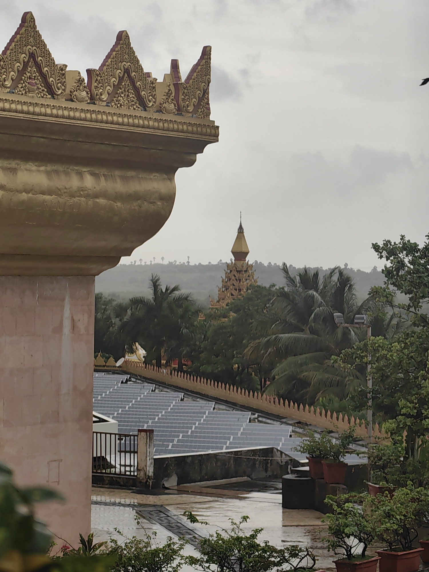 The image depicts a view of the Global Vipassana Pagoda in Gorai, Mumbai, India, on a cloudy day. The foreground features the ornate golden architectural details of a structure, likely part of the pagoda complex. Below this, rows of solar panels are visible on a rooftop, suggesting a modern integration of technology with traditional architecture. Beyond the solar panels, lush green trees and palm trees create a dense canopy. In the distance, the distinctive tiered golden spire of the Global Vipassana Pagoda rises above the treeline. The sky is overcast, indicating a potentially humid or rainy weather condition, typical of Mumbai's climate. A single dark bird is captured in flight against the sky. The overall scene conveys a sense of peace and spiritual grandeur, juxtaposed with functional modern elements.