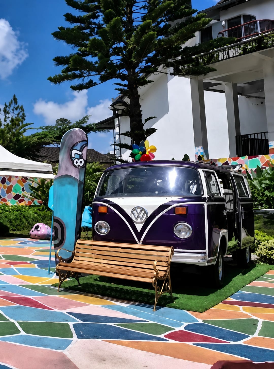 In Berastagi, Indonesia, a vibrant scene unfolds beneath a bright blue sky with scattered white clouds. The foreground is dominated by a colorful, geometric-patterned pavement in shades of blue, pink, green, yellow, and red. A wooden bench with ornate gold legs sits on a patch of artificial green grass, positioned in front of a purple and white Volkswagen Type 2 van. The van's doors are open, and a colorful pinwheel adorns its roof. To the left of the van, a tall, slender blue surfboard with a graphic design featuring the number "87" stands upright. In the background, a large tree with dark green foliage towers over the scene, with a white building featuring balconies and dark railings visible behind it. Lush green foliage surrounds the van and extends into the mid-ground, where a pink, polka-dotted object is partially visible.