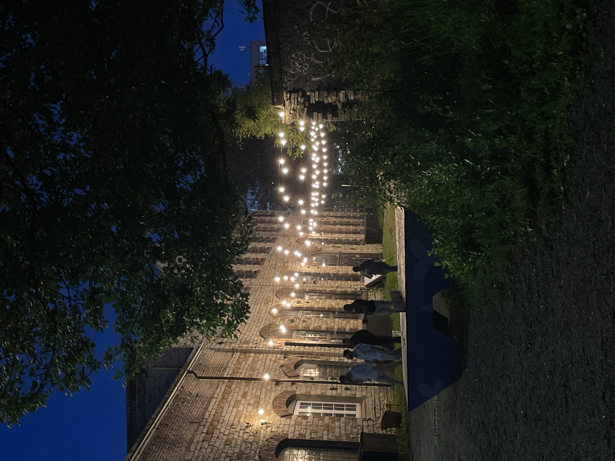 This photograph, taken at dusk in Tallinn, Estonia, captures an outdoor seating area adjacent to a weathered brick building. The building, characterized by arched windows and rustic brickwork, is illuminated by a string of warm-toned Edison bulbs that stretch across the scene. Several individuals are visible near the building, some appearing to be walking while others are gathered. In the foreground, a dark blue, angular seating structure, possibly a bench or a table, is positioned on a gravelly surface. Lush green foliage and grass surround the seating area and the lower portion of a stone wall to the right. The sky above is a deep twilight blue. The overall impression is one of a relaxed evening gathering in an urban, yet natural, setting.