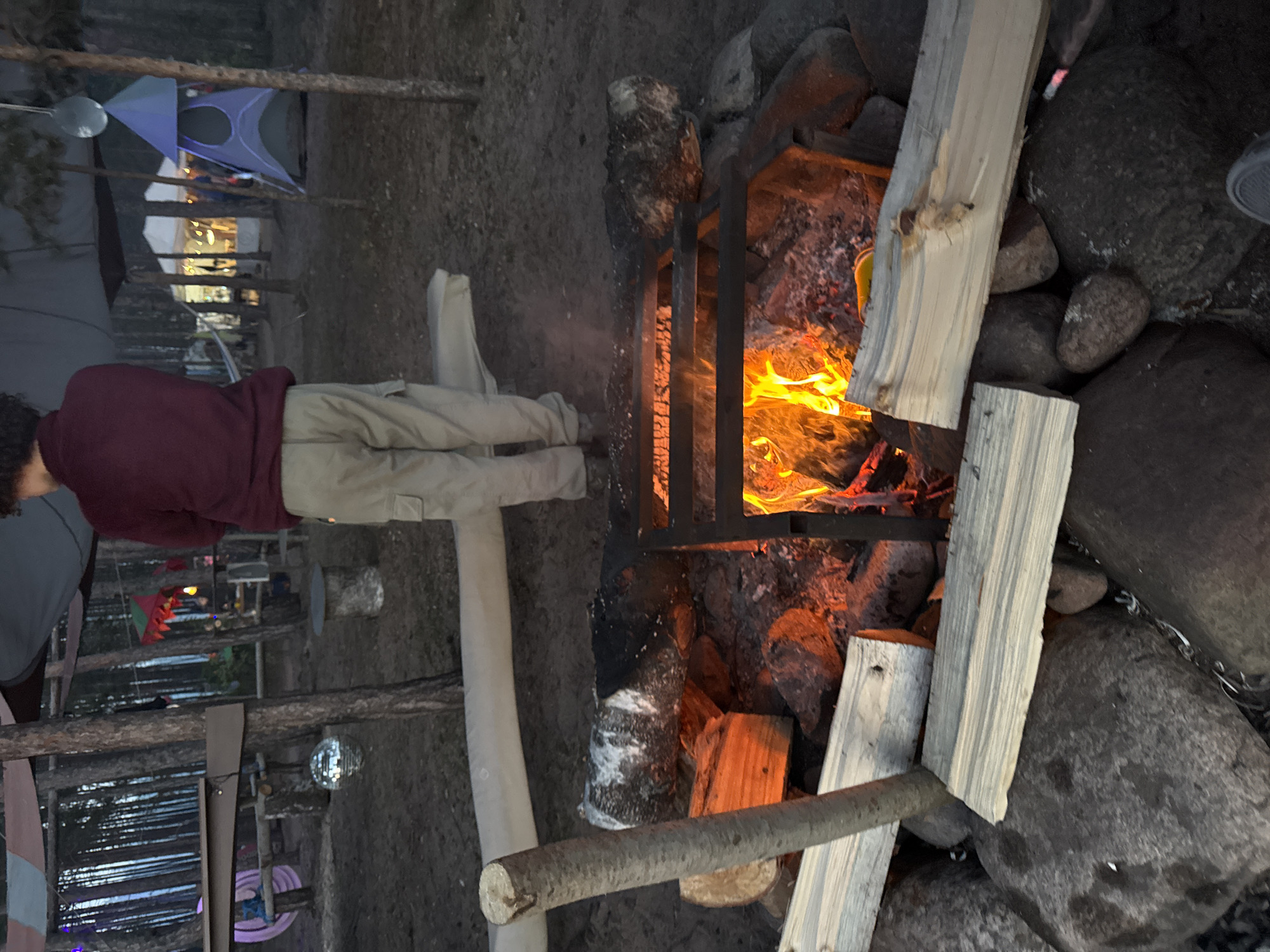 A campfire burns within a black metal fire pit surrounded by large rocks. Pieces of light-colored wood lean against the rocks and fire pit. To the left, a person wearing a maroon shirt and khaki pants stands facing the fire. In the background, tents and decorative items are visible amidst trees. The location is potentially in or near Luzhki, Russia.