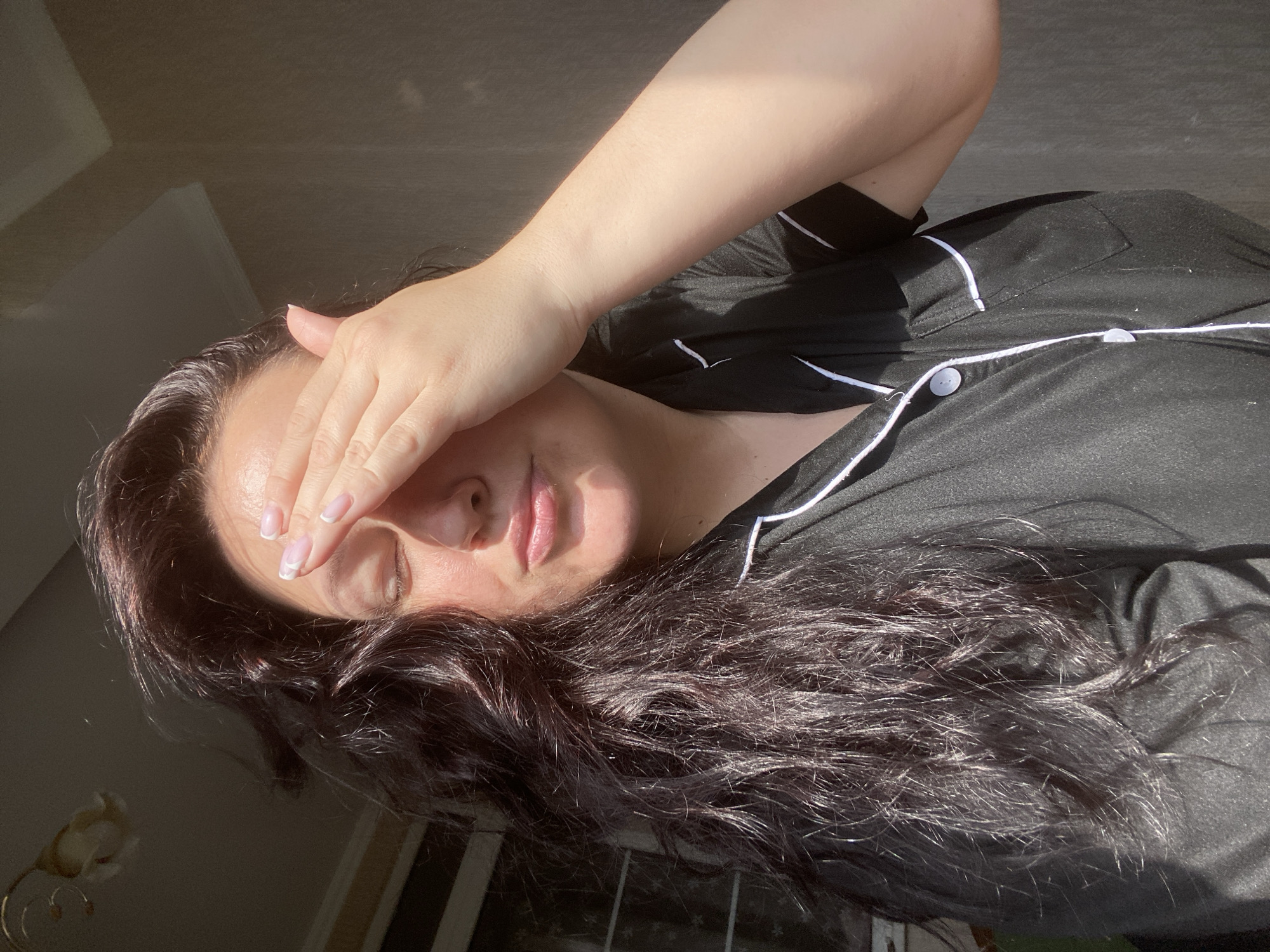 A medium shot depicts a woman with long, dark hair, wearing black pajamas with white piping, shielding her face with her right hand from bright sunlight. Her nails are manicured with a French tip. She appears to be indoors, given the presence of an unseen wall or window frame on the left. The background is nondescript, suggesting a domestic interior. No evidence indicates the location is Sochi, Russia.