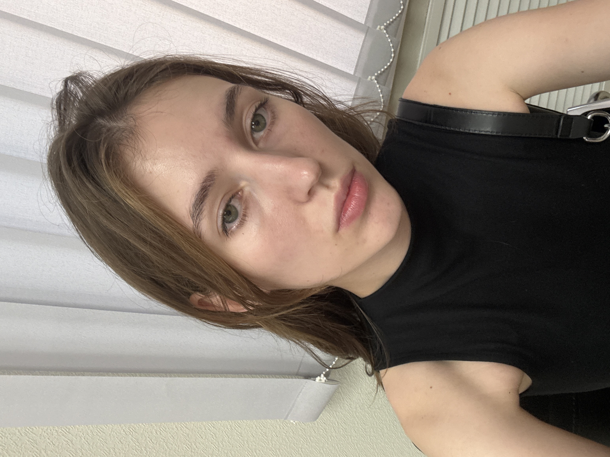 A young woman with light brown hair and green eyes is looking directly at the camera in a selfie. She is wearing a black sleeveless top. The background consists of vertical blinds and a textured wall. The lighting appears to be natural, coming from the right. There is no identifiable information to suggest the image was taken in Perm, Russia.